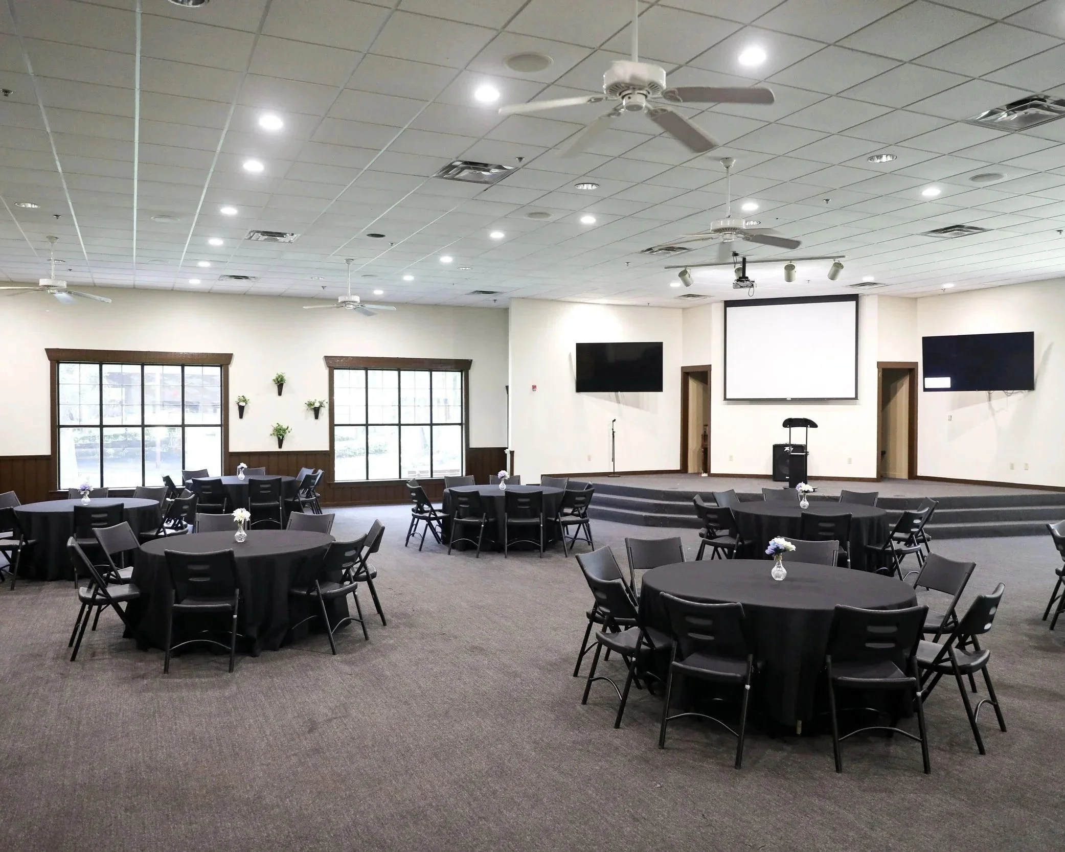 Large conference room with round tables covered with black tablecloths, each with a small flower arrangement. The room has multiple ceiling fans, recessed lighting, and large windows with wooden trim. Two large screens are mounted on the front wall.