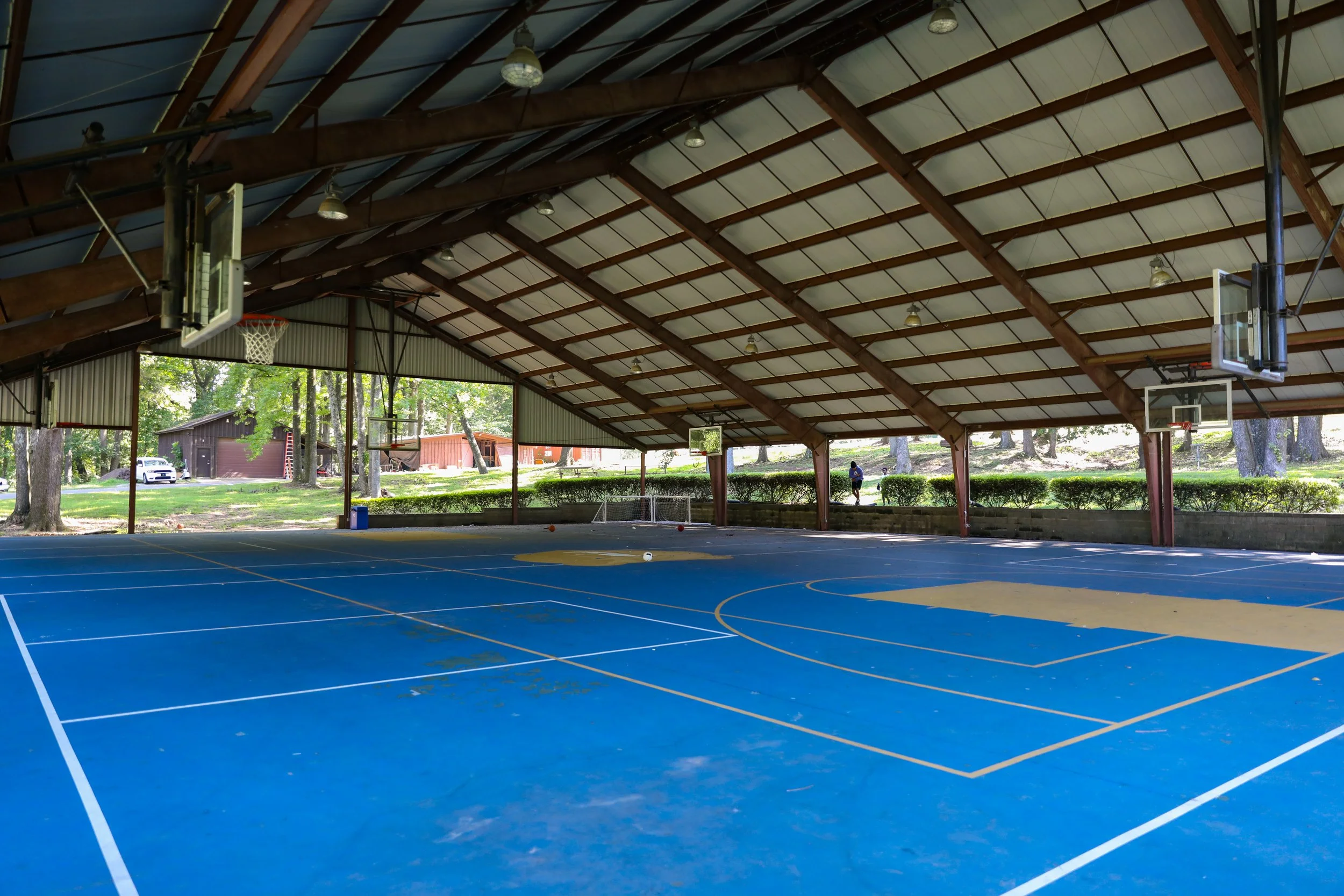 Indoor basketball court with blue flooring, white court lines, and multiple basketball hoops, surrounded by large open sides showing trees and a building outside.