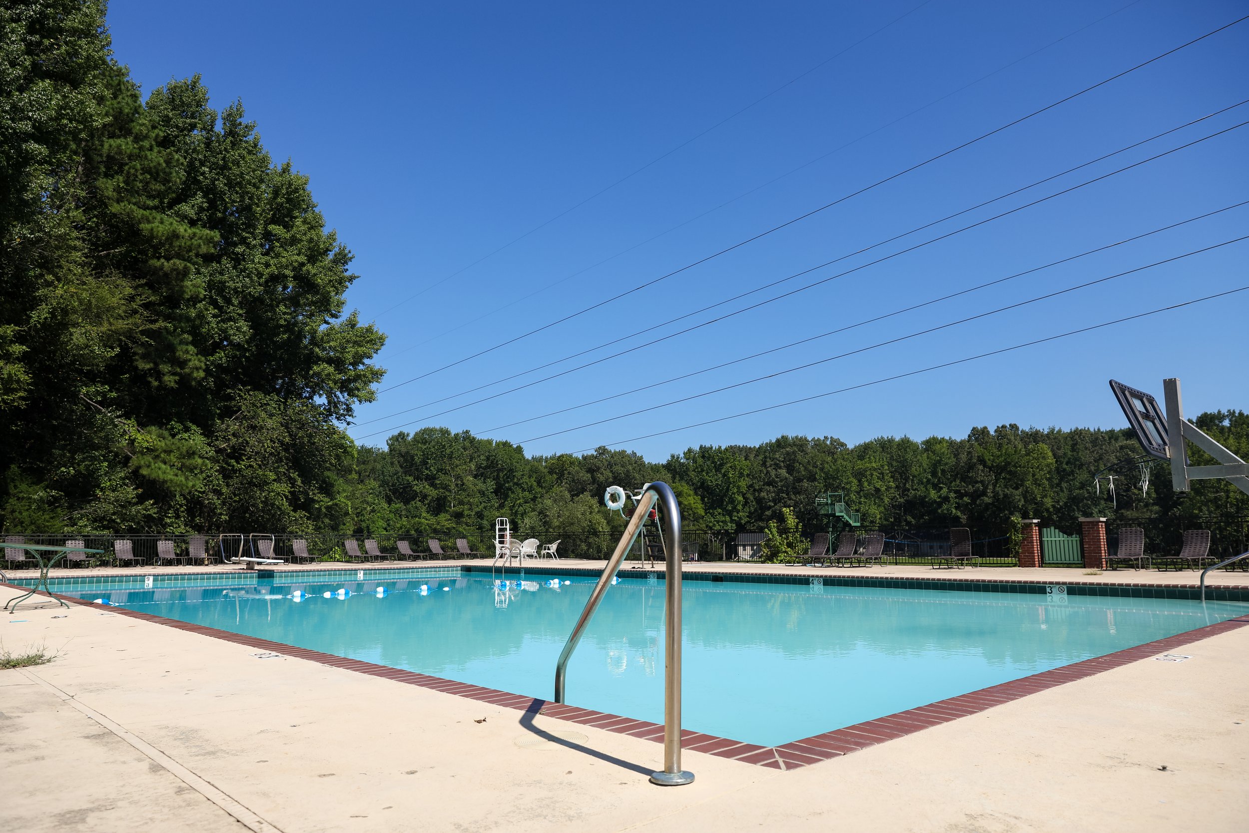An outdoor swimming pool with empty lounge chairs, surrounded by trees and a clear blue sky.