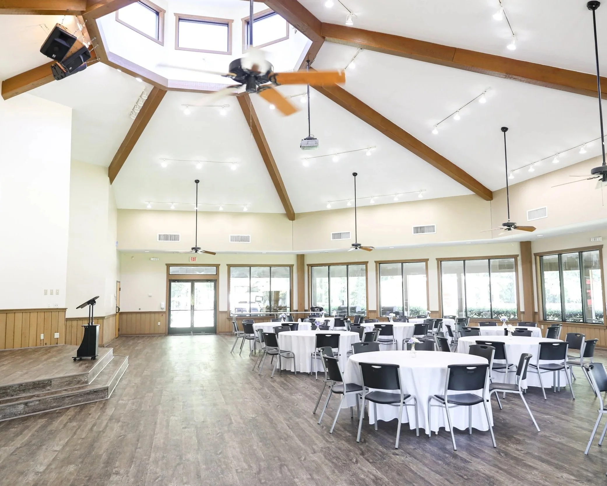 A spacious banquet hall or event space with round tables covered in white tablecloths and black chairs, wooden flooring, high ceiling with wooden beams and multiple hanging ceiling fans, large windows allowing natural light, and a small stage with steps at the front.