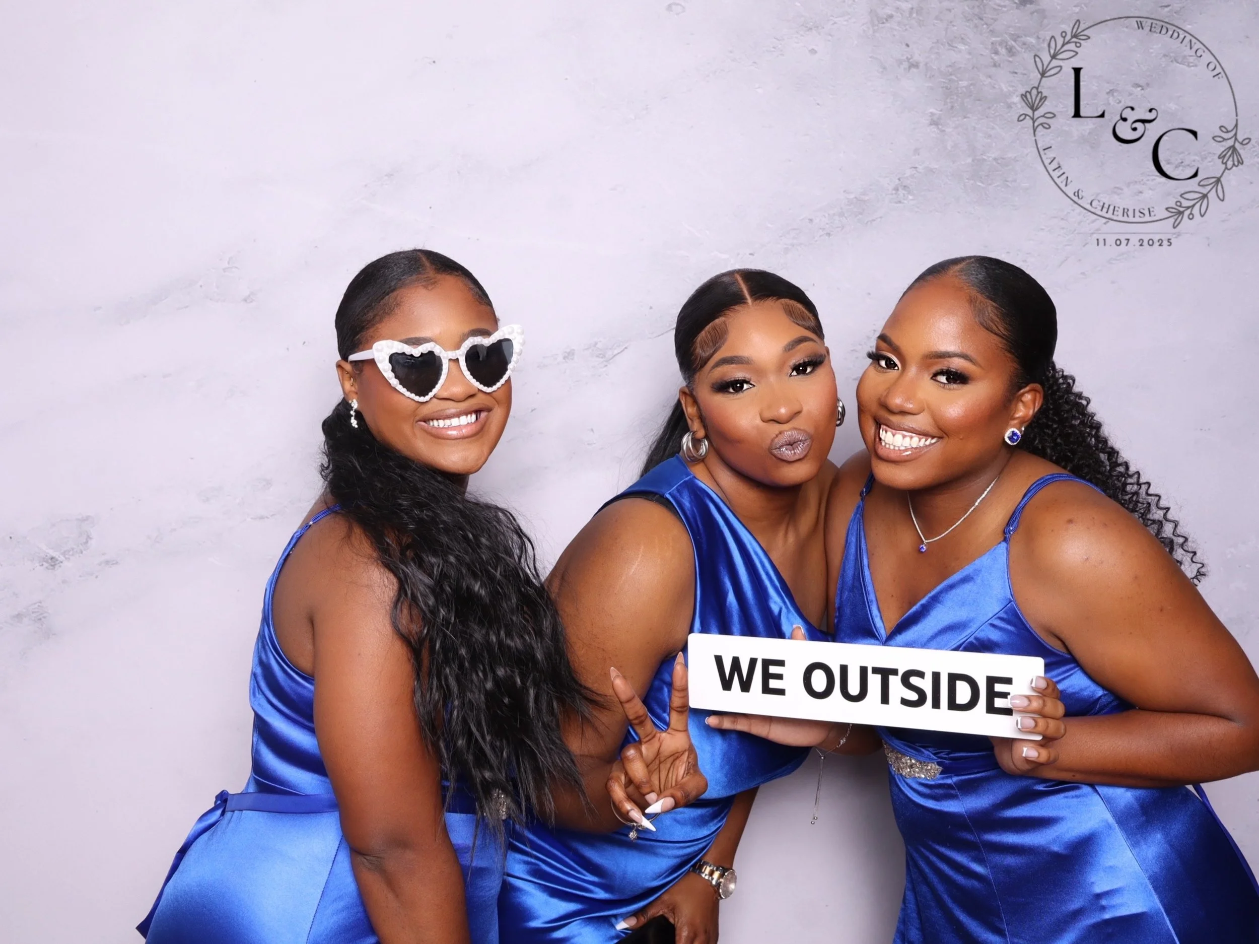 Three women dressed in blue satin dresses posing together at a wedding, one holding a sign that says "WE OUTSIDE". One woman is wearing heart-shaped sunglasses, and they are all smiling.