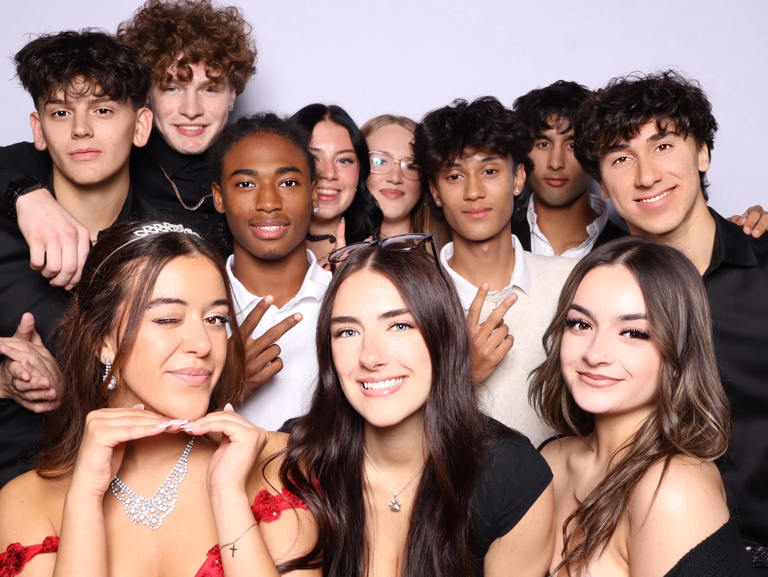 Group of teenagers and young adults taking a group photo, smiling, making peace signs, and posing playfully against a plain background.