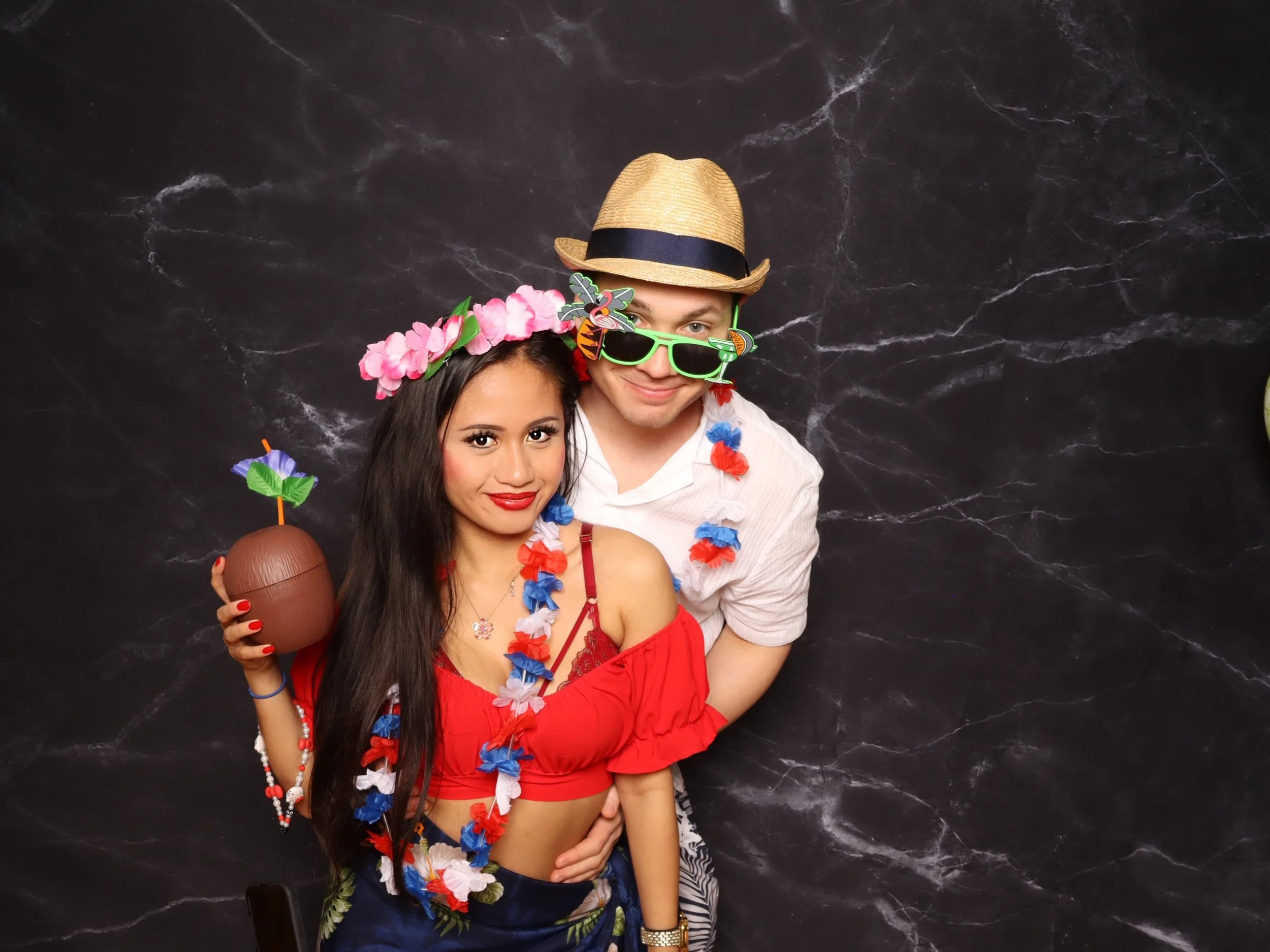 A young woman in a red dress, wearing a pink flower lei and holding a coconut drink, stands next to a young man in a white shirt, with a straw hat, green sunglasses, and a blue, red, and white lei. They are posing against a black marble background.
