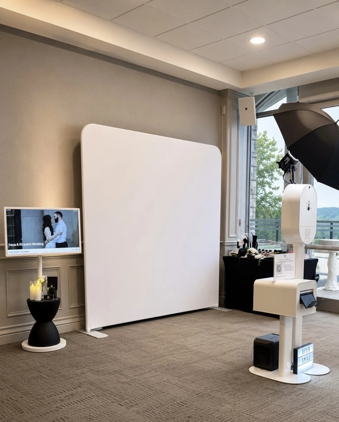 Photo booth setup inside a room with a blank photo backdrop, a screen displaying a wedding photo, and various props for taking pictures, near a large window with trees outside.