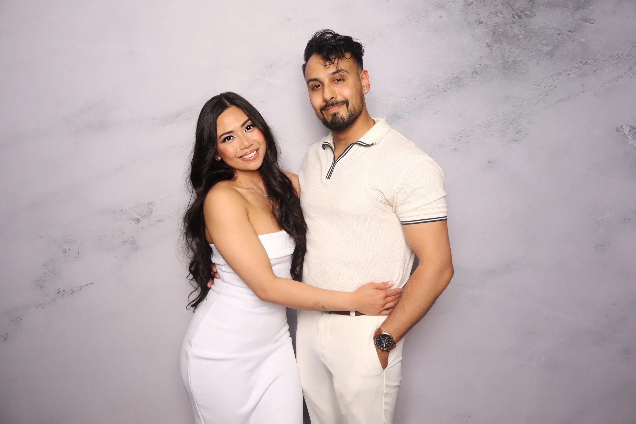 A smiling woman in a white strapless dress and a man in a cream polo shirt and white pants standing close together against a gray wall, with the woman's arm around the man's waist and the man's arm around the woman's back.