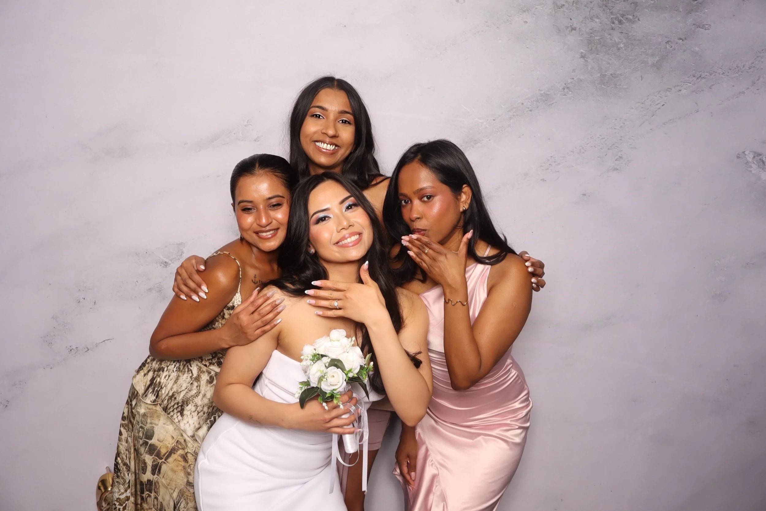 Group of five women in formal dresses posing together against a plain background, with one holding a bouquet of white flowers.