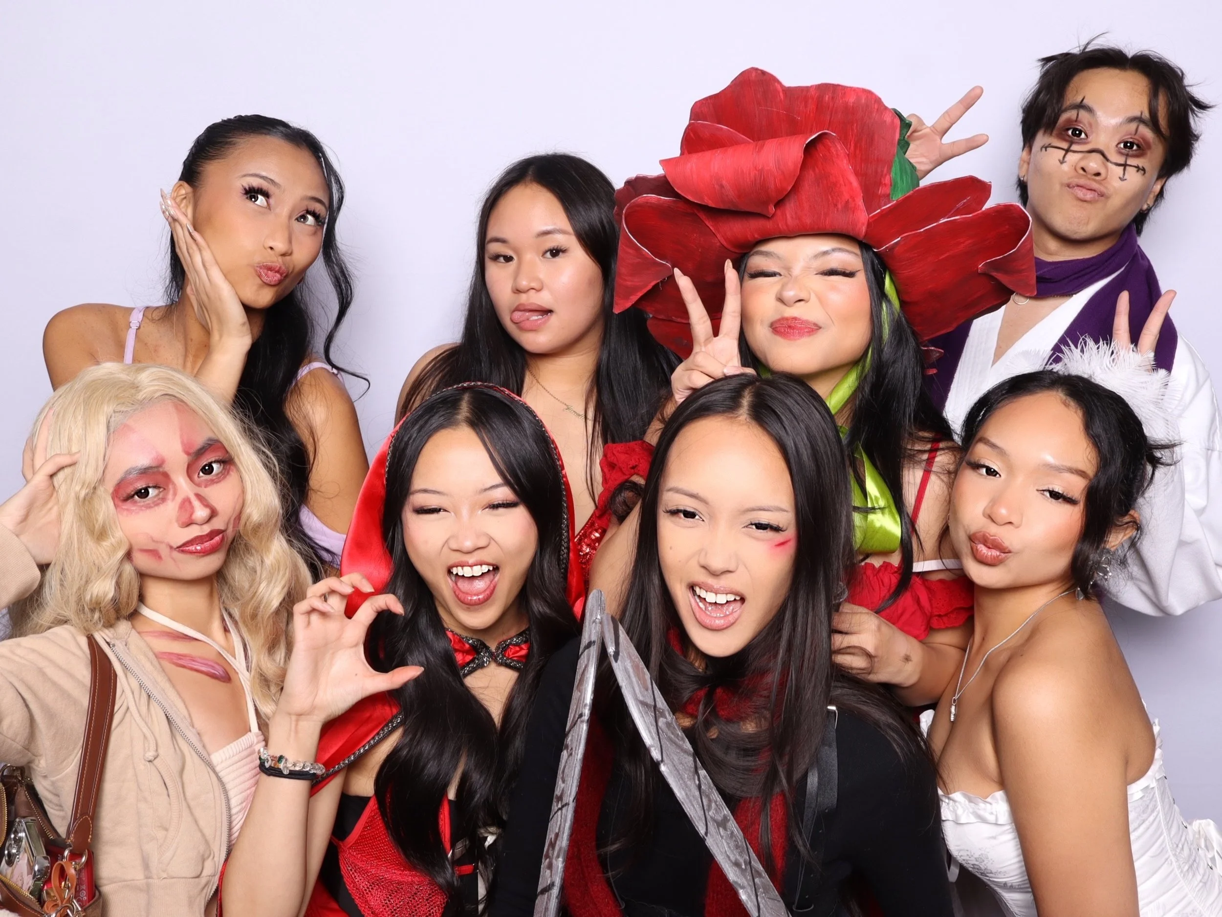 Group of nine young adults in costumes posing for a photo, some with makeup and accessories, making playful facial expressions and gestures.