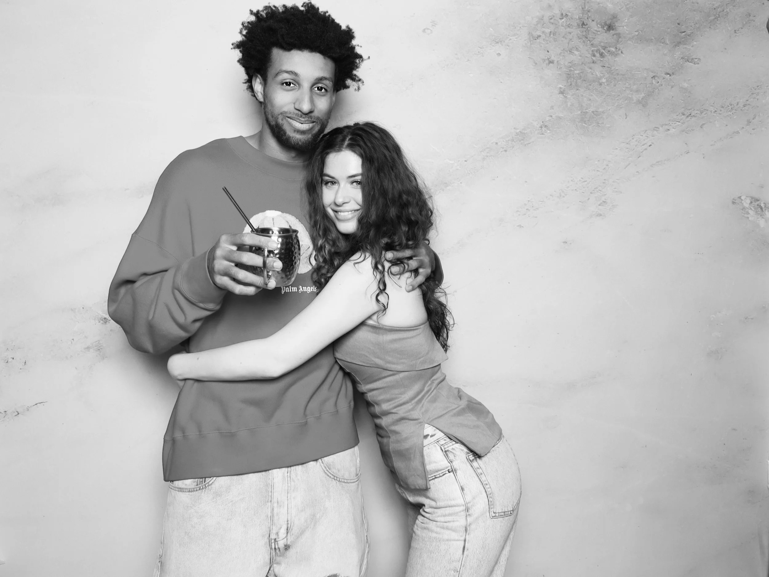 A man and woman posing together, with the man holding a cup with a straw and a lemon slice. They are smiling and embracing each other against a plain wall background.