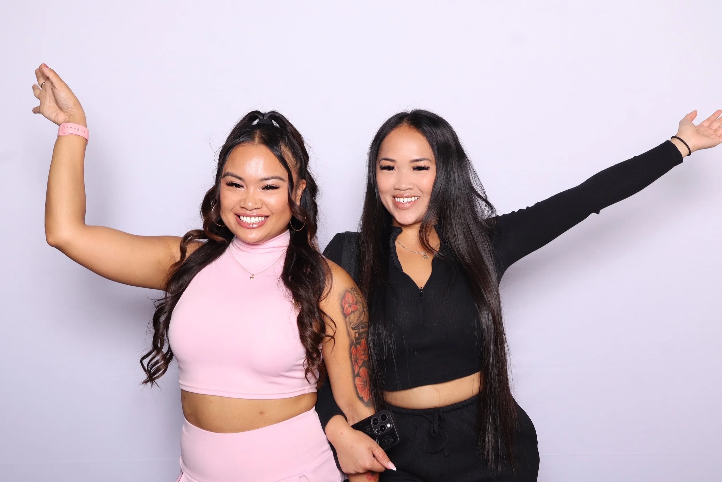 Two women smiling and posing with arms raised against a plain white background.