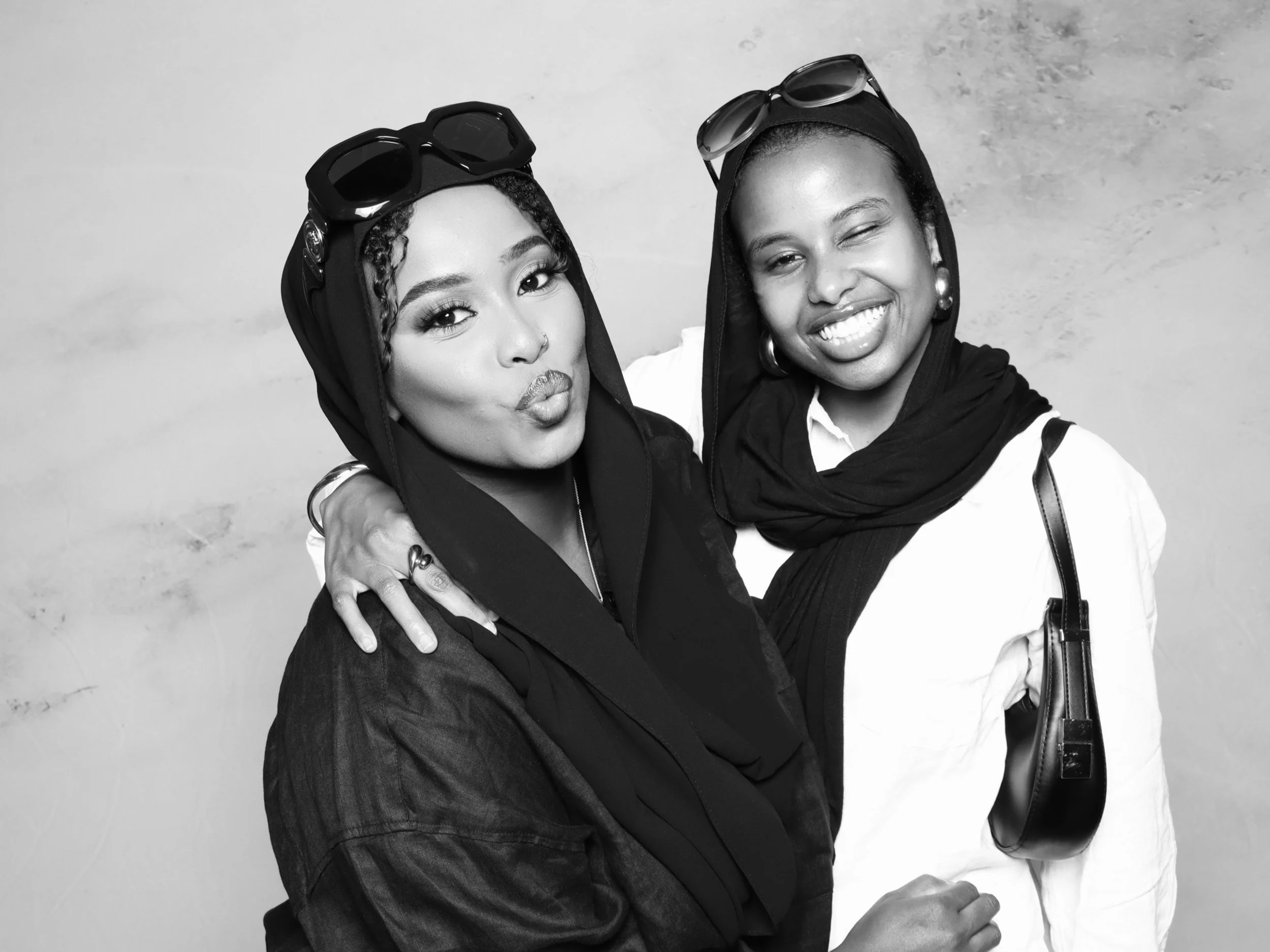 Two women wearing hijabs smiling and hugging in front of a plain background, with one winking and the other blowing a kiss.