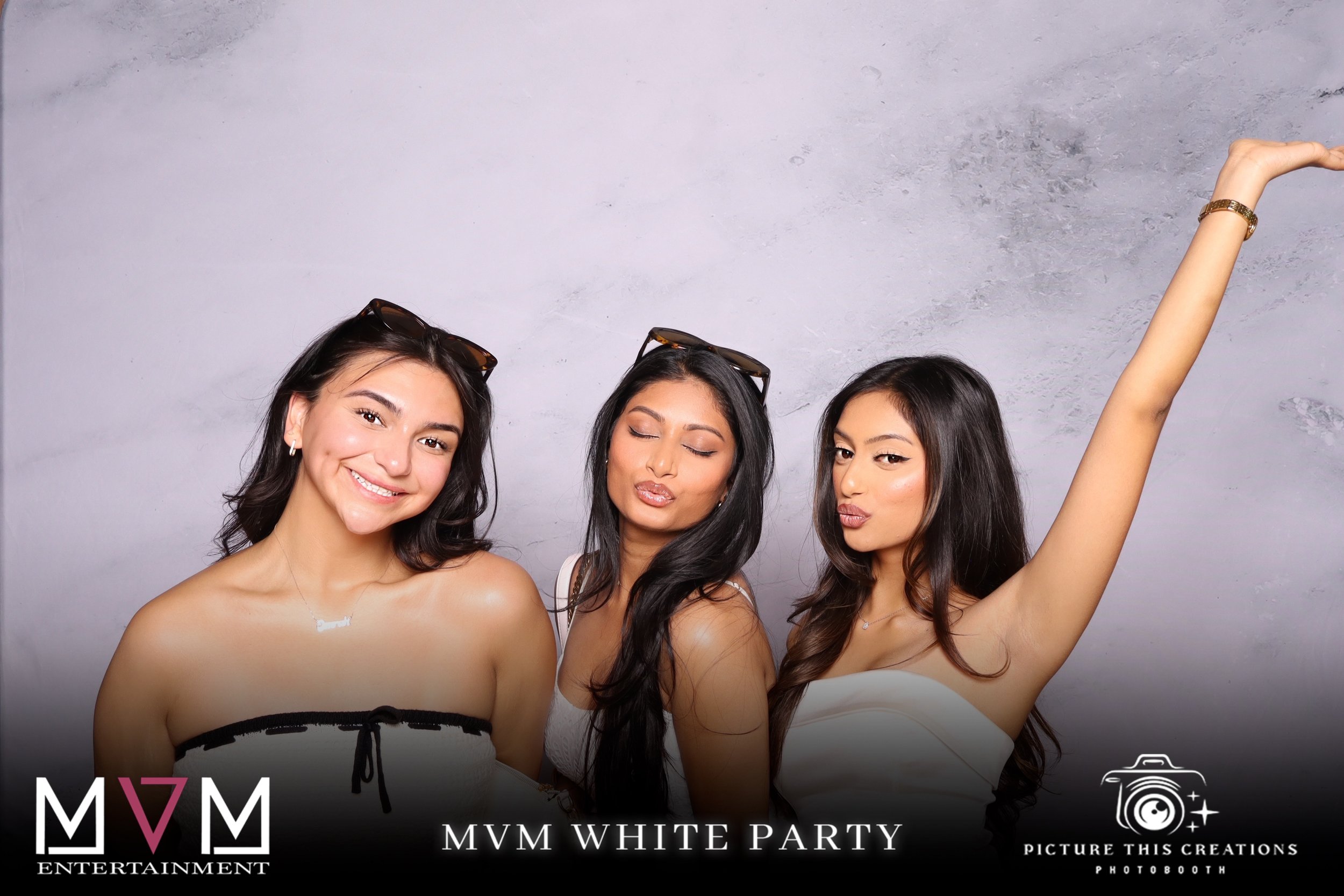 Three young women posing for a photo at a white party with gray background. They are dressed in white tops, wearing sunglasses on their heads, and making playful facial expressions. The woman on the left is smiling, the one in the middle is making a 