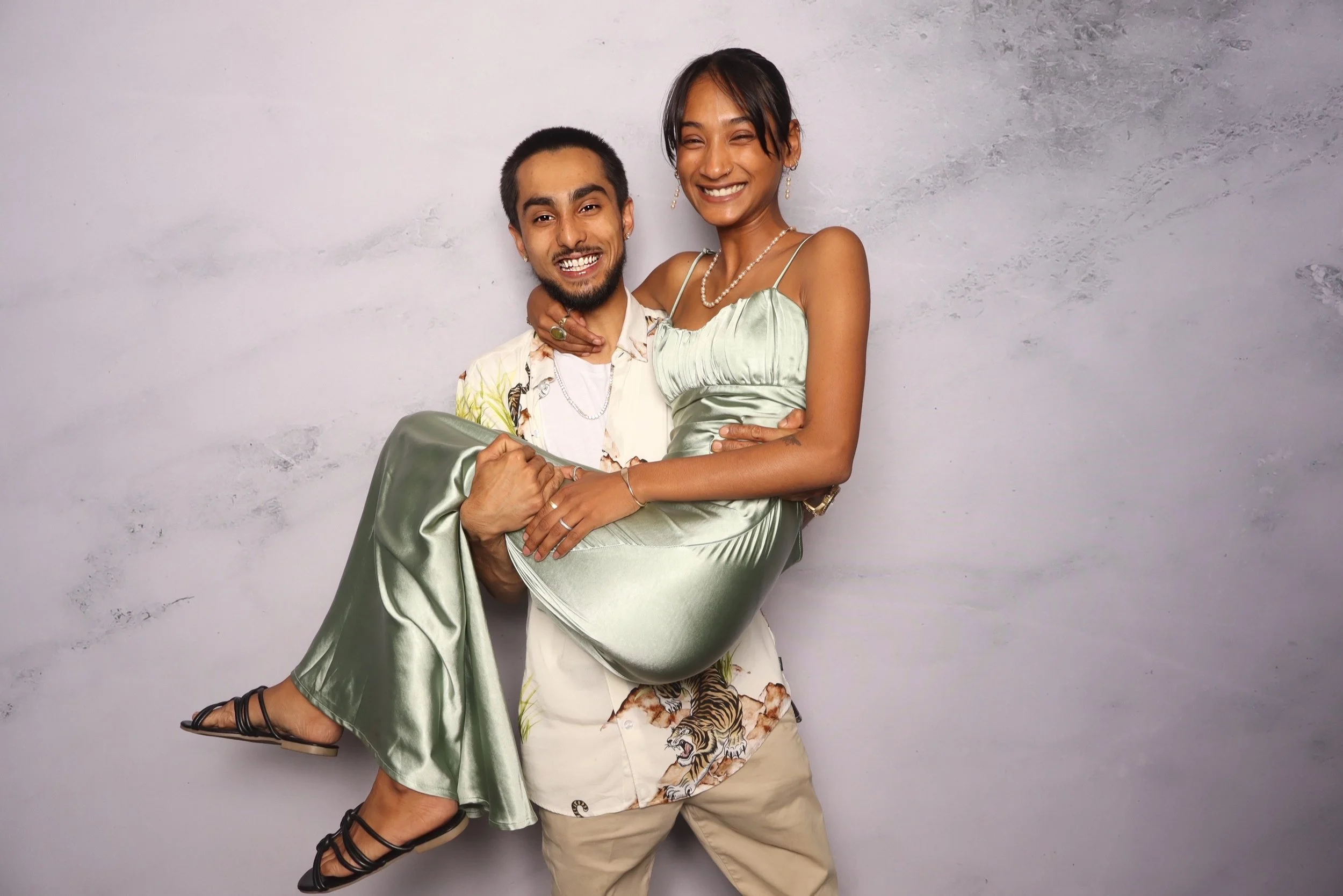 A man holding a woman in his arms, both smiling and wearing stylish clothing, standing against a plain gray background.
