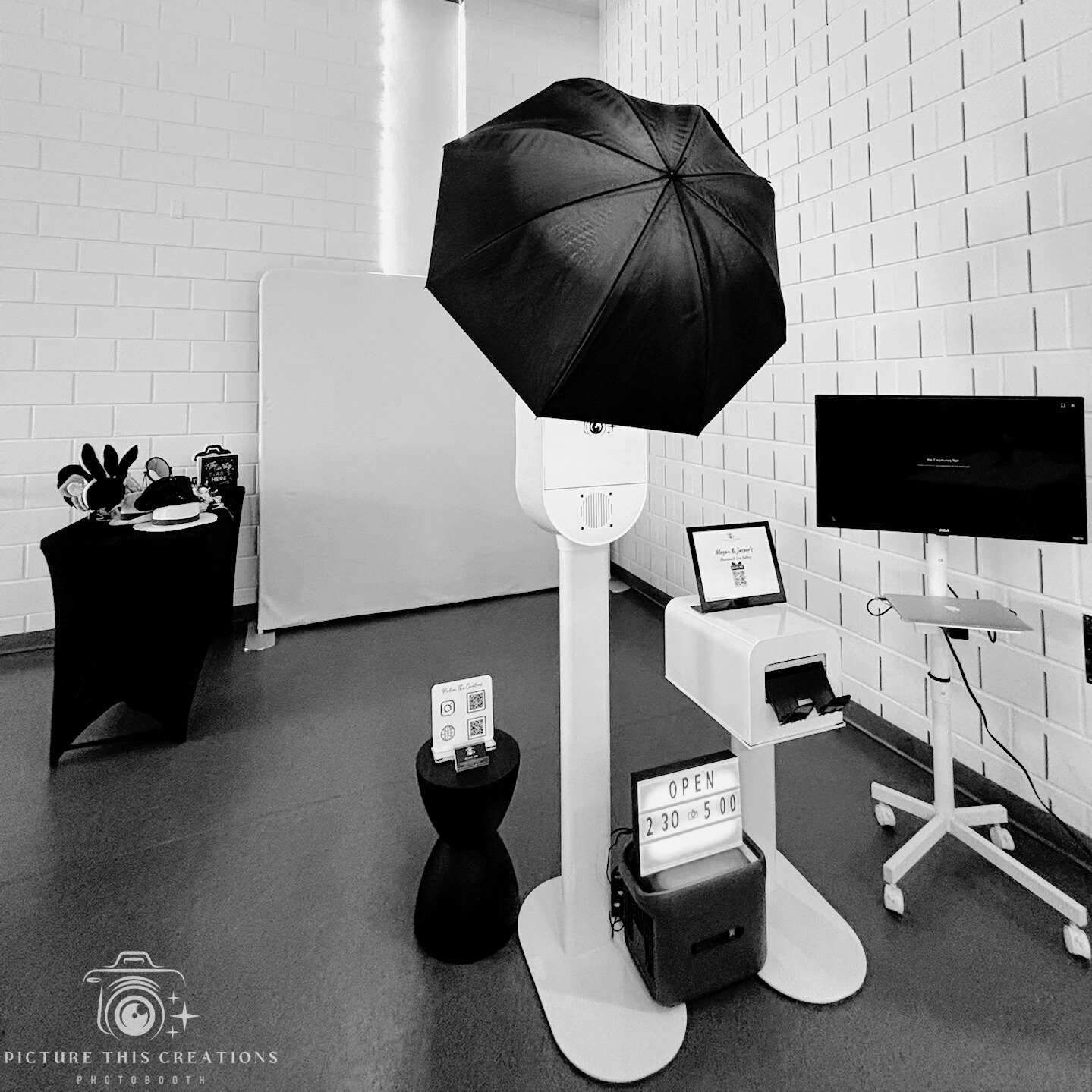 A photo booth setup with a large black umbrella, a small table with decorative items, a digital sign, and a screen, all in a room with white brick walls.