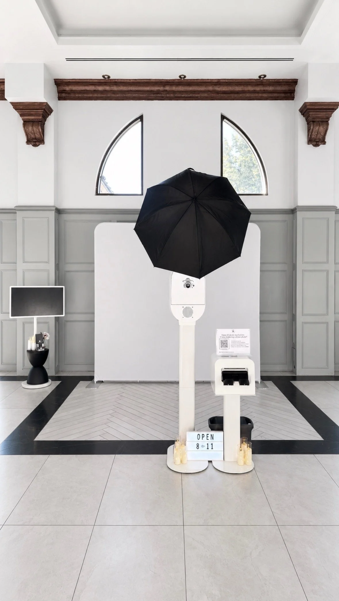 Photo of a photo booth with a black umbrella over a camera on a white stand, candles at the base, and a sign indicating the hours of operation from 8 to 11. The background includes a white wall with decorative crown molding, arched windows, and a sma