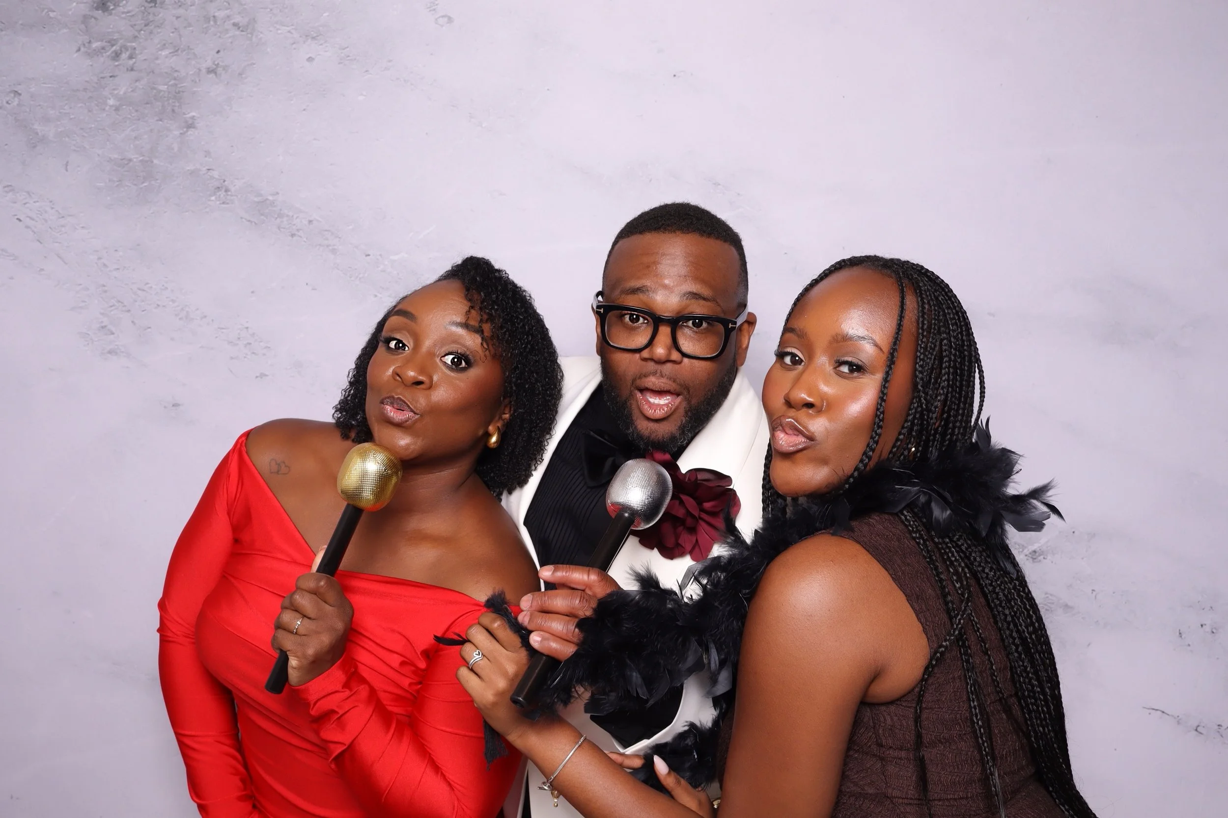 Three people posing with microphones against a gray background, dressed in elegant attire, with the two women wearing feather boas and the man wearing glasses and a tuxedo.