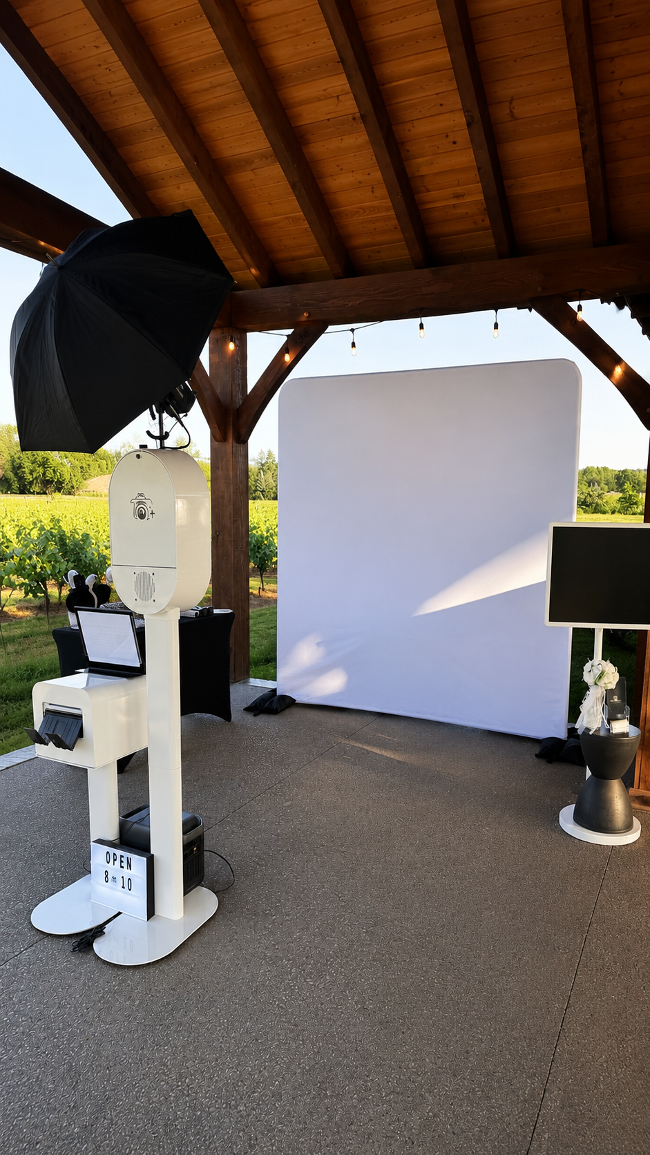 Photo of a photo booth setup outdoors under a wooden pavilion with string lights. Includes a white backdrop, photo booth printer, camera equipment, a TV, and various accessories, with a green vineyard or farm in the background.