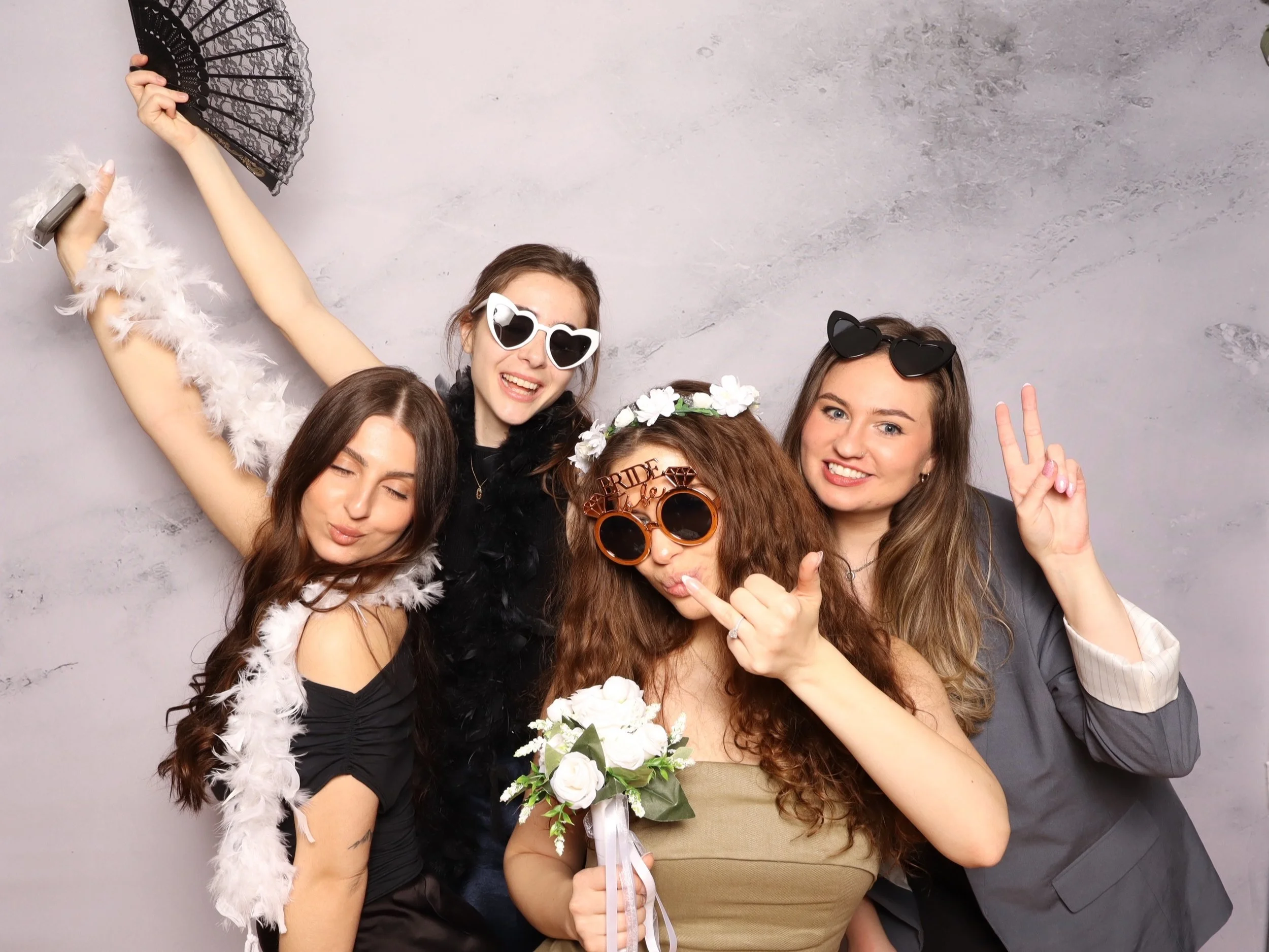 Group of five women celebrating, wearing funny glasses and accessories, one woman holding a bouquet, some making peace signs, cheerful expressions.