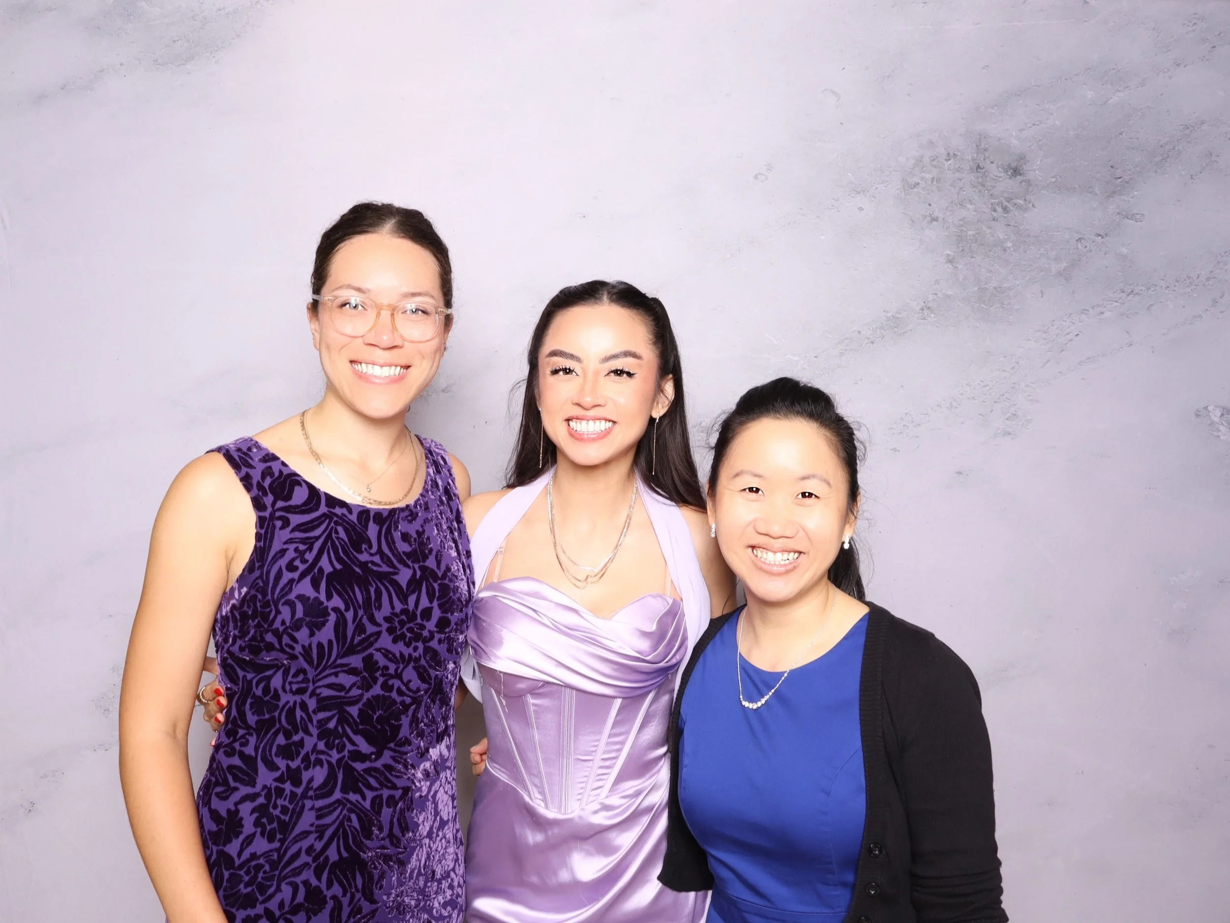 Three women smiling in front of a gray wall, dressed in colorful outfits.