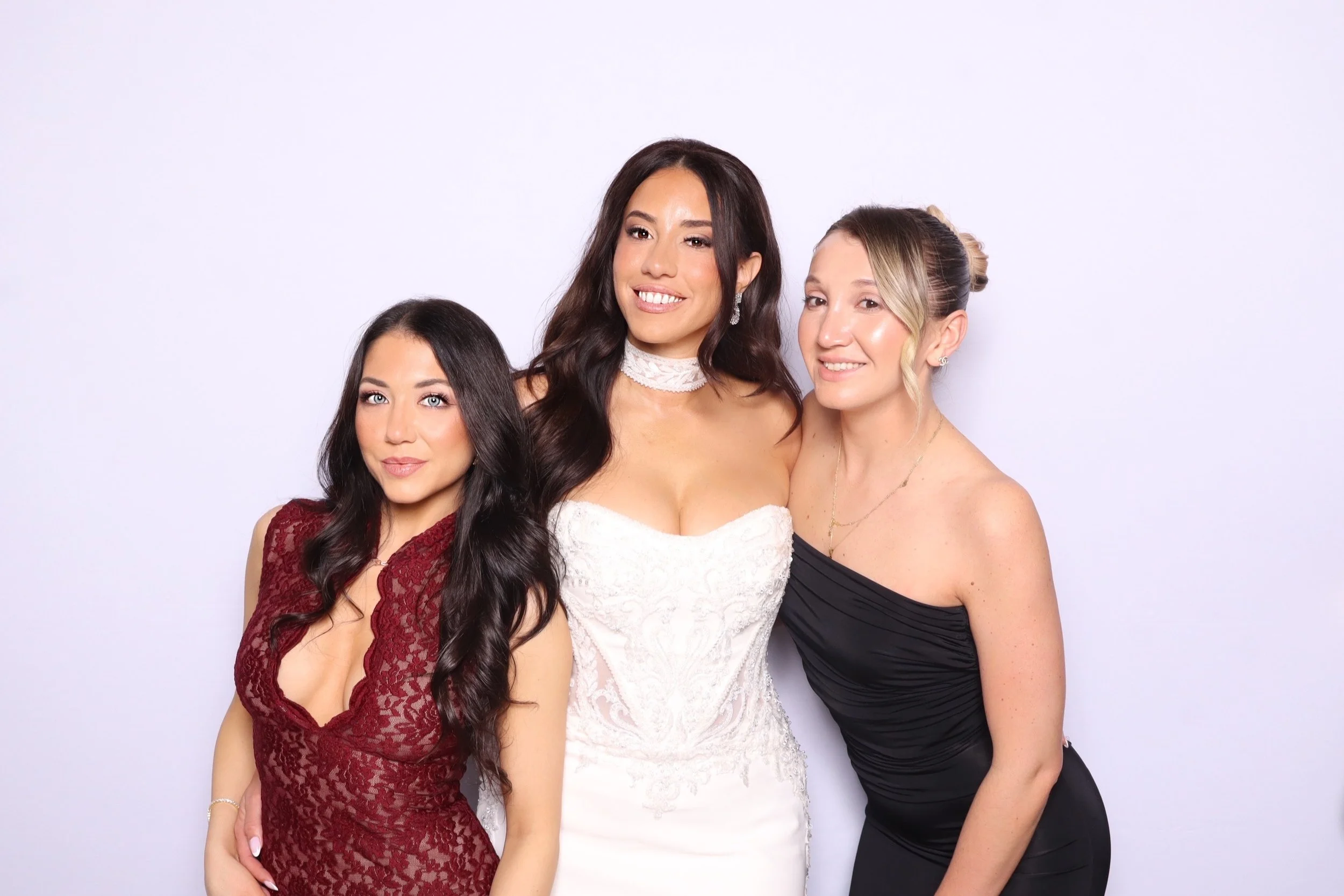 Three women dressed elegantly, standing close together against a plain white background, smiling at the camera.