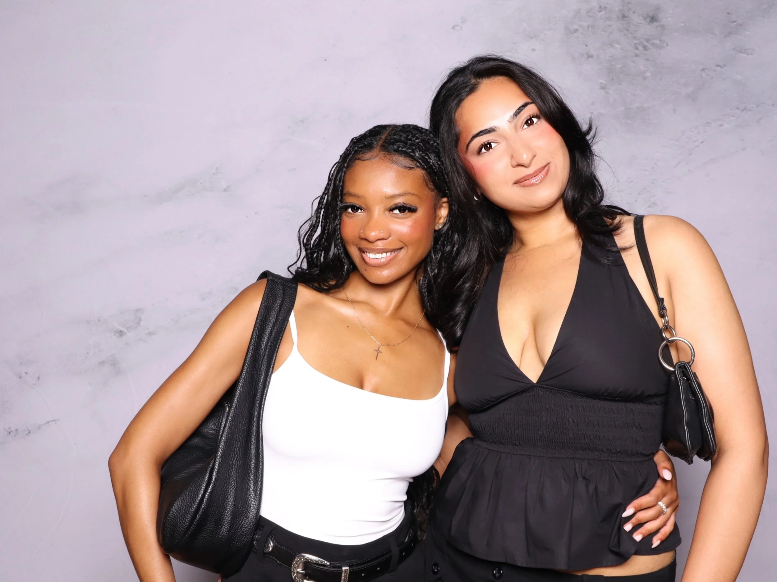 Two women smiling, standing close together against a gray wall. One has dark curly hair and a white tank top, the other has wavy black hair and a black sleeveless top.