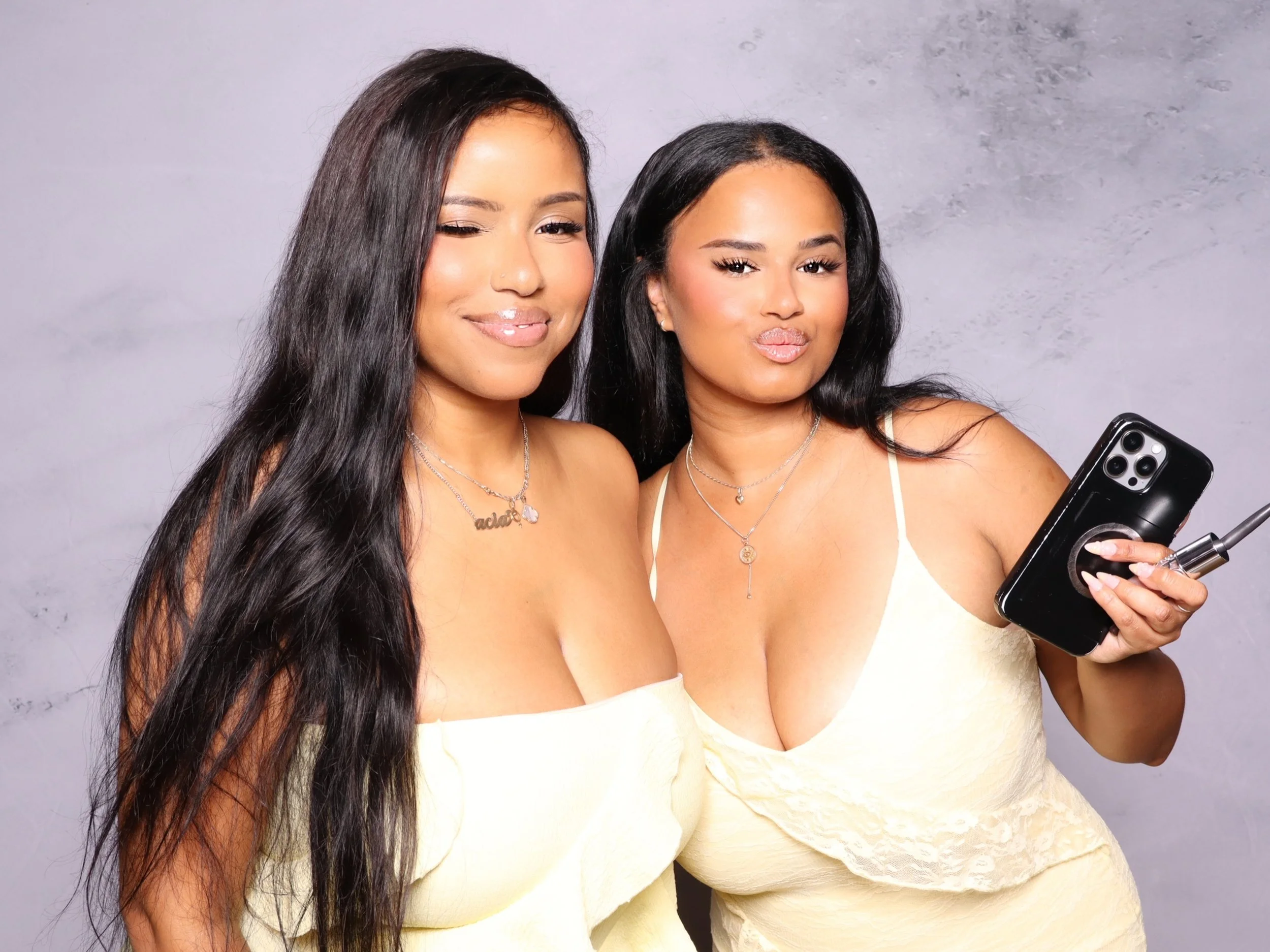 Two women with long dark hair taking a selfie with a smartphone, both wearing light-colored dresses and necklaces, standing against a plain light-colored background.