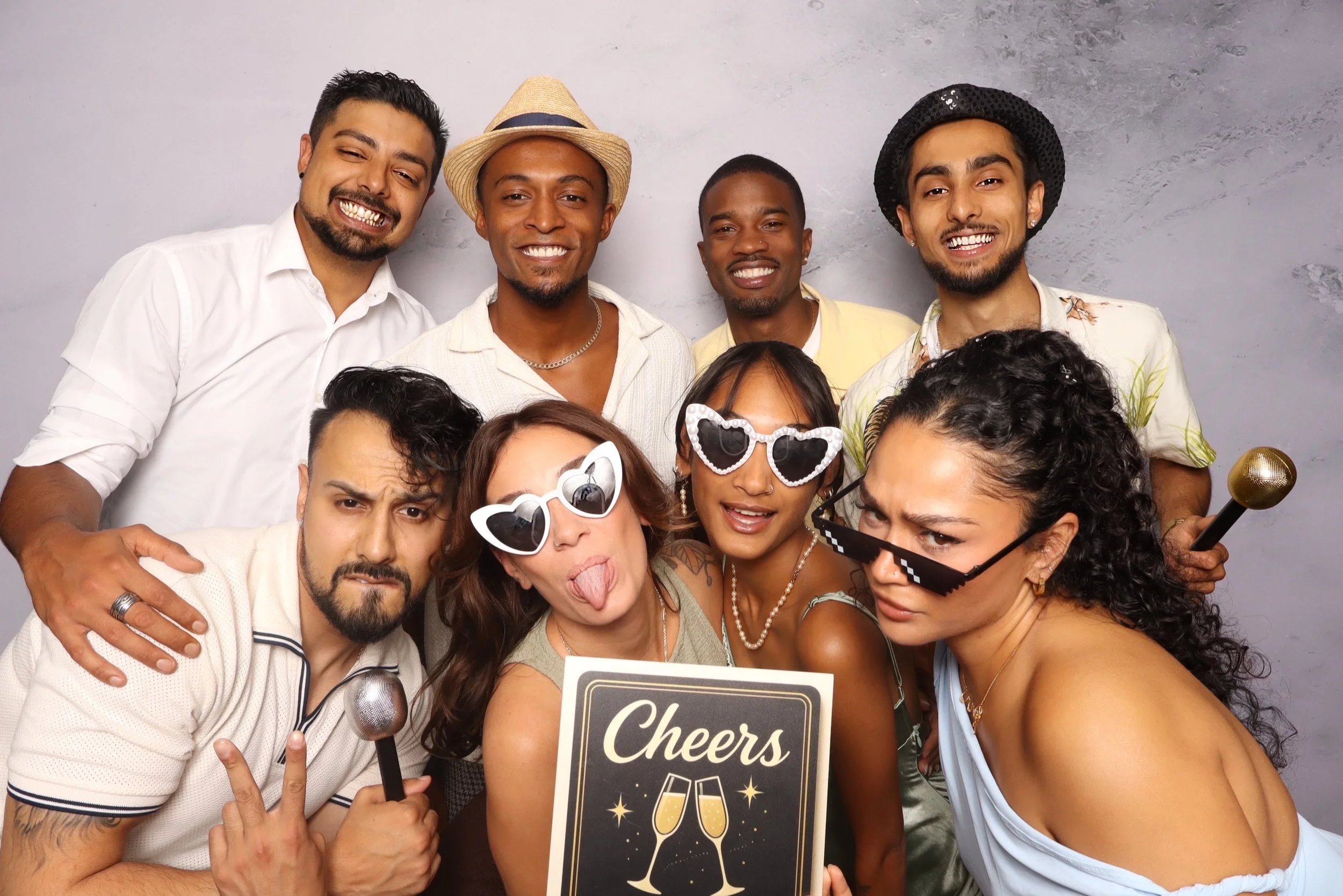Group of people celebrating with party props, sunglasses, drinks, and a cheer sign, smiling and posing for the camera.