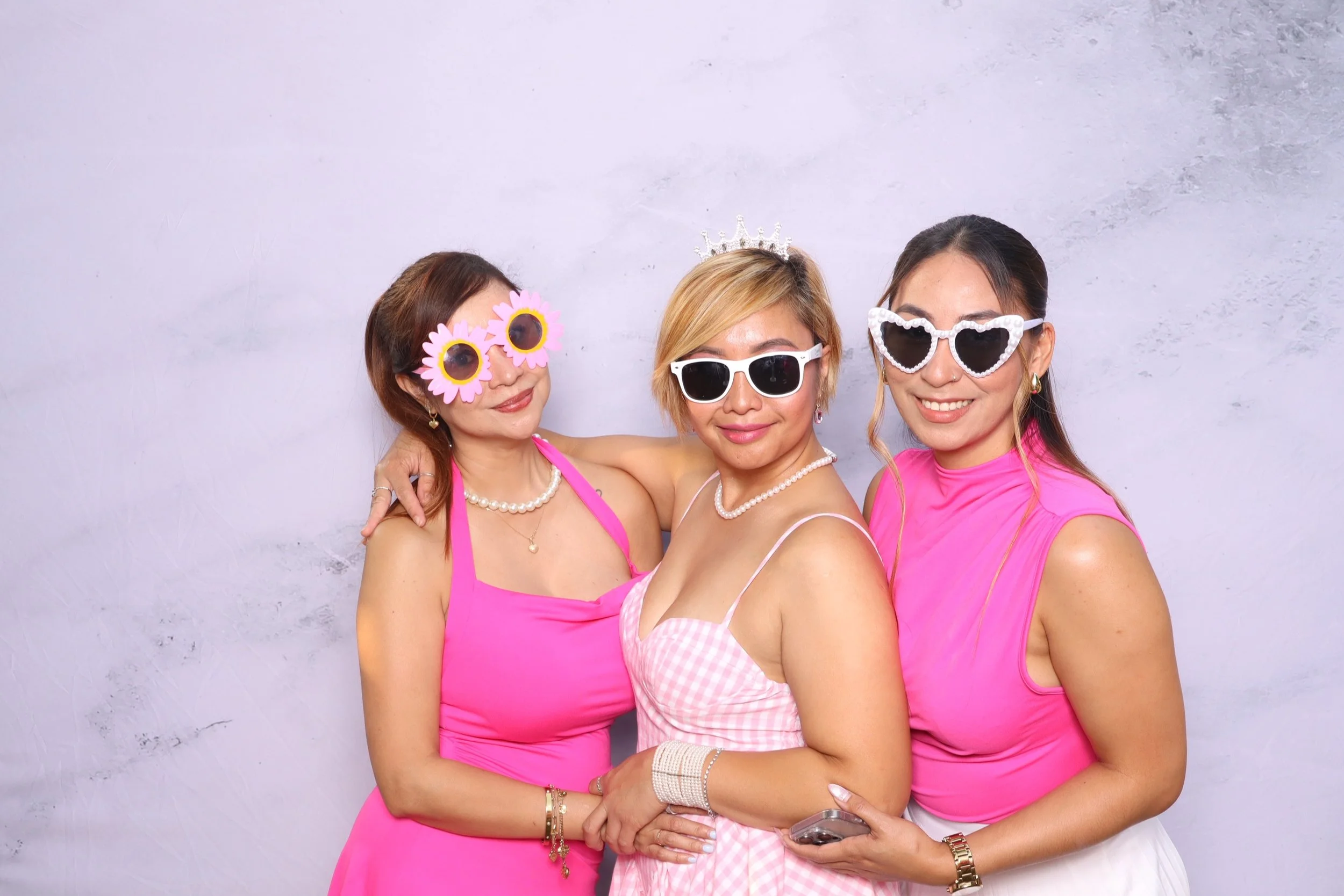 Three women dressed in pink wearing fun sunglasses and accessories, smiling at the camera, standing against a plain light purple background.