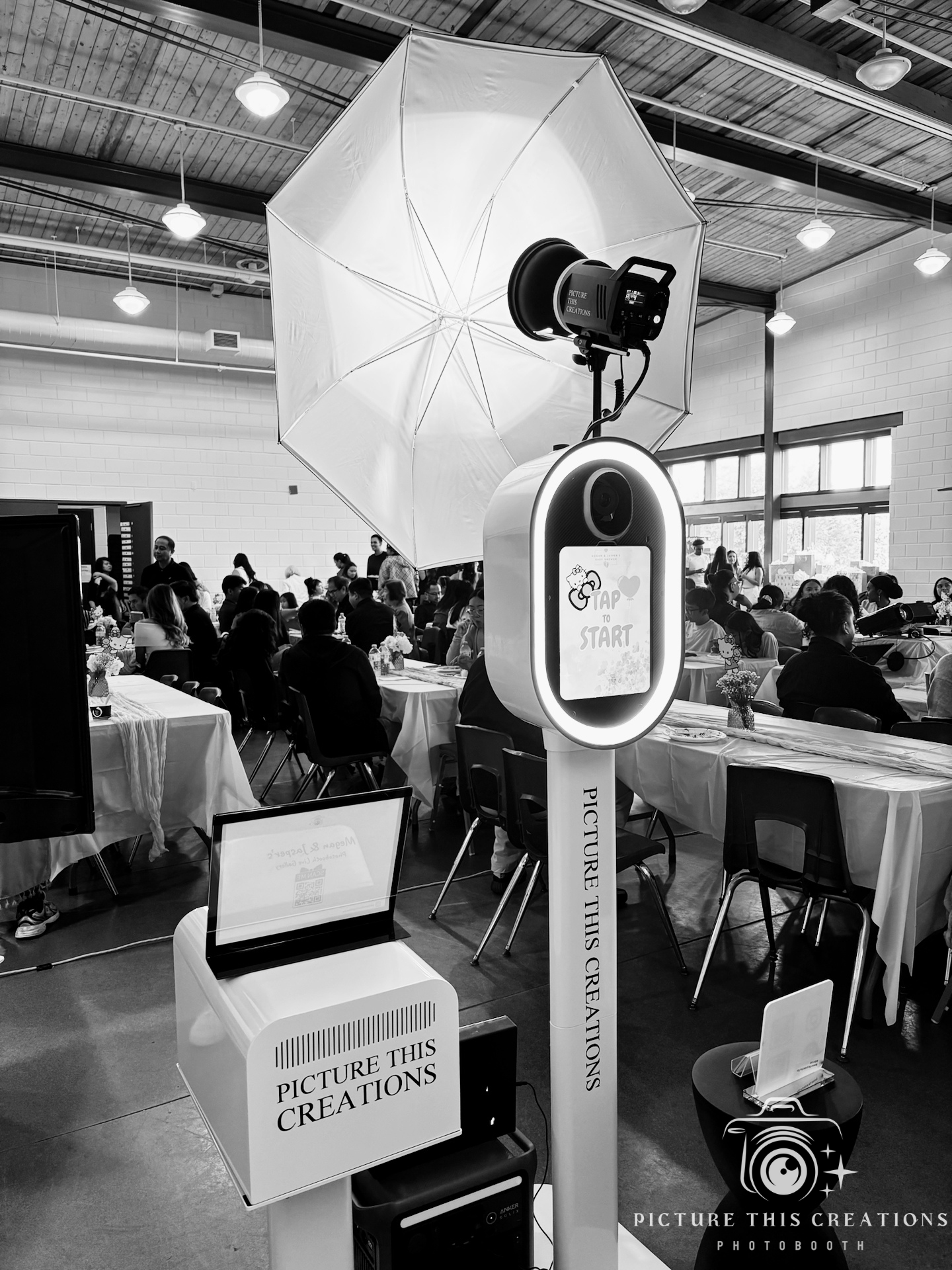 A photo booth setup with a large umbrella light, camera, and a digital screen displaying 'Tap to Start' inside a busy event space with people seated at decorated tables.