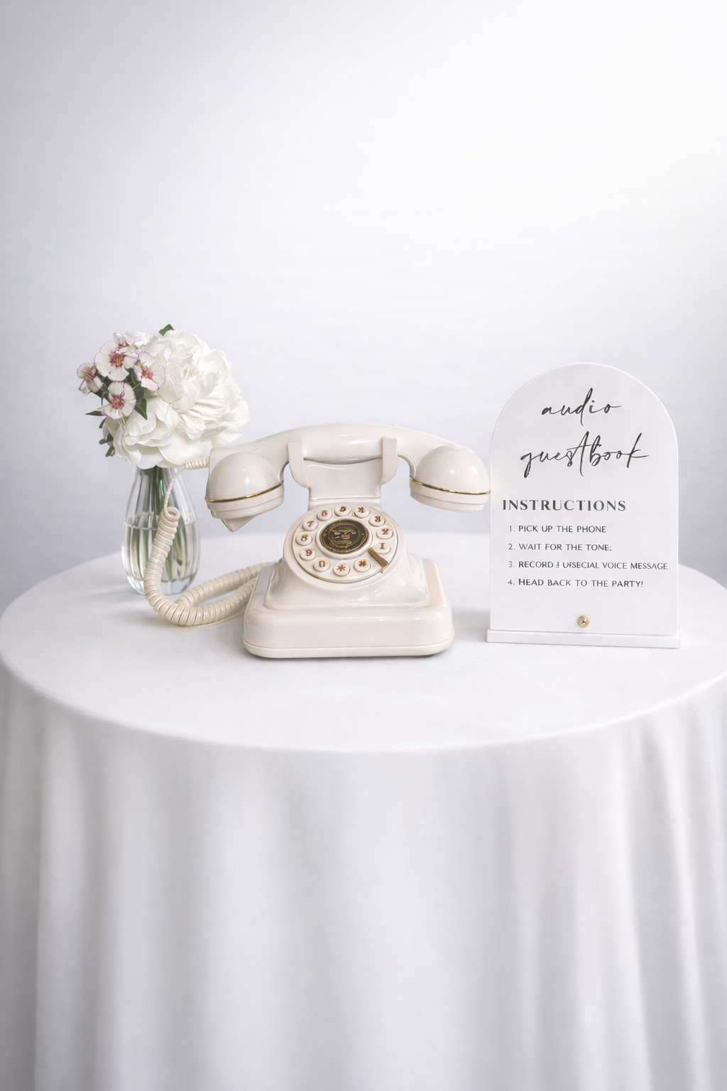 A vintage white rotary telephone on a white round table with a vase of white flowers, and a sign with instructions for an audio guestbook.