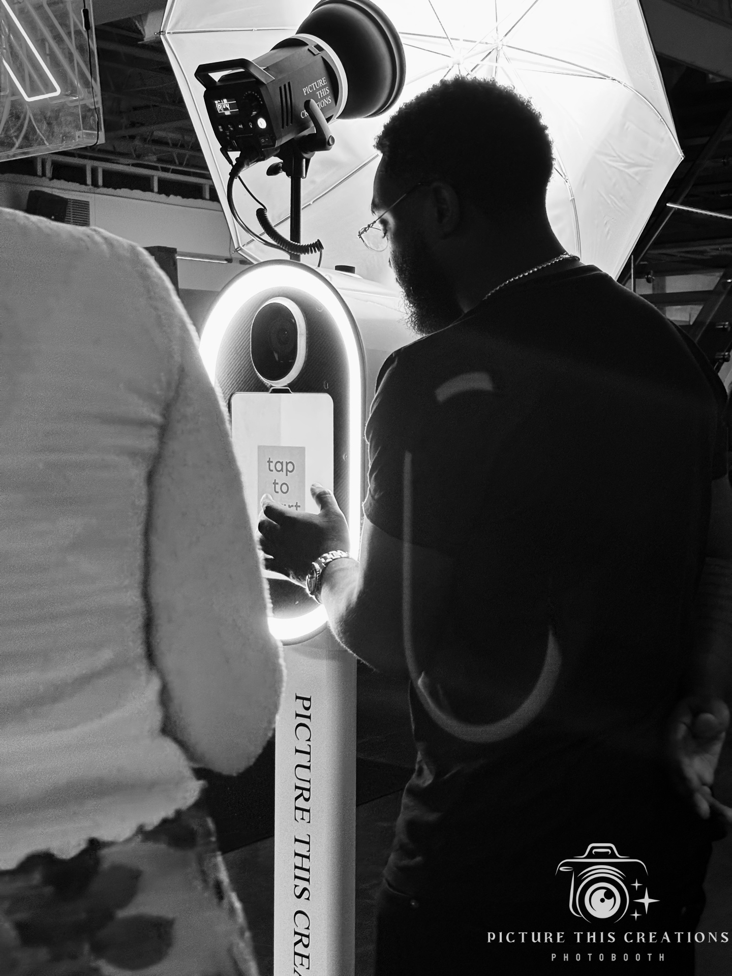 A man with glasses and a beard using a smartphone at a photo booth, with a large circular camera and studio lighting equipment behind him. The photo booth has a vertical sign that says 'PICTURE THIS CREATIONS' and a smaller sign on the camera with th
