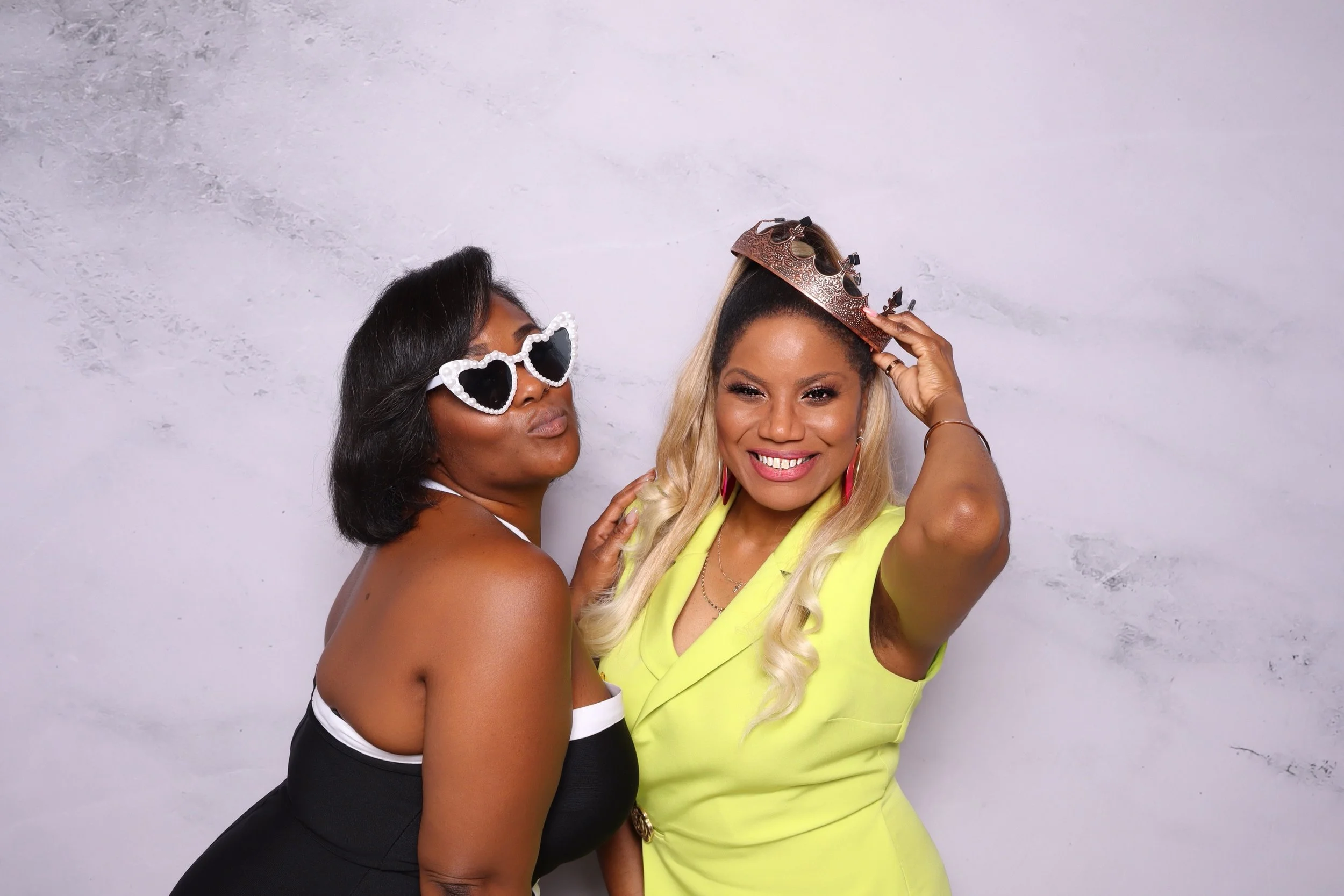 Two women celebrating, one wearing heart-shaped sunglasses and the other holding a crown on her head, both smiling and posing in front of a plain background.