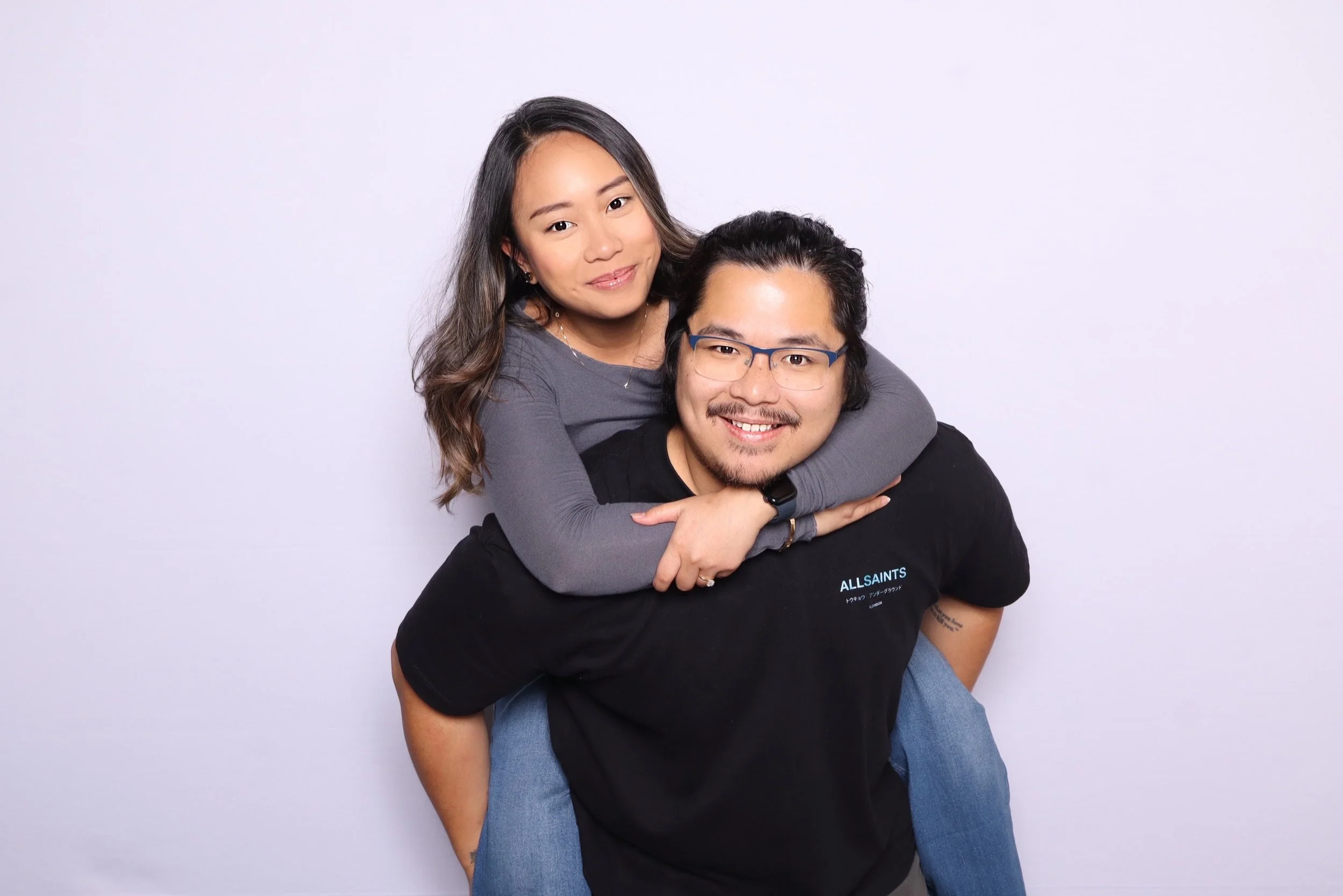 A smiling woman with long dark hair giving a piggyback ride to a smiling man with glasses and a mustache, against a plain white background.