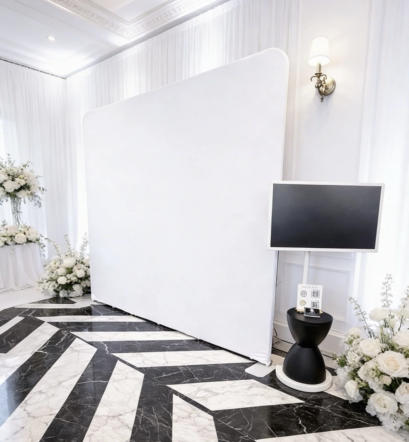 Interior of a room with black and white marble flooring, a white folding partition, a flat-screen TV on a stand, a floral arrangement on the left, a wall sconce light, and a small black table with informational signs.