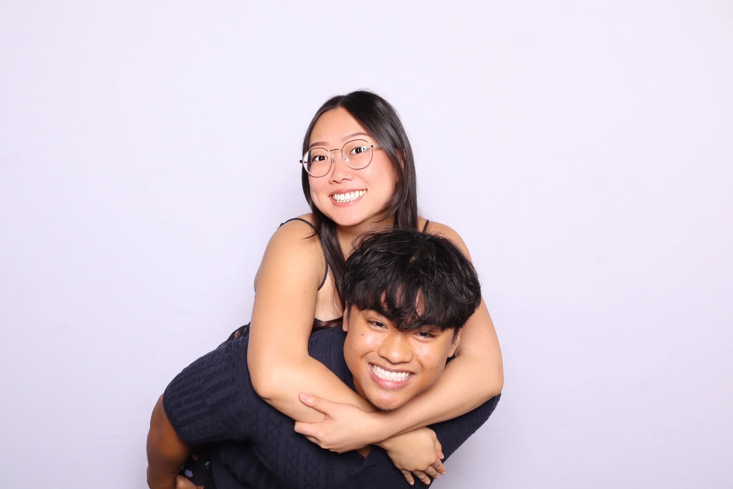 A smiling young woman with glasses and long dark hair on a white background, being piggybacked by a young man with dark hair, also smiling.