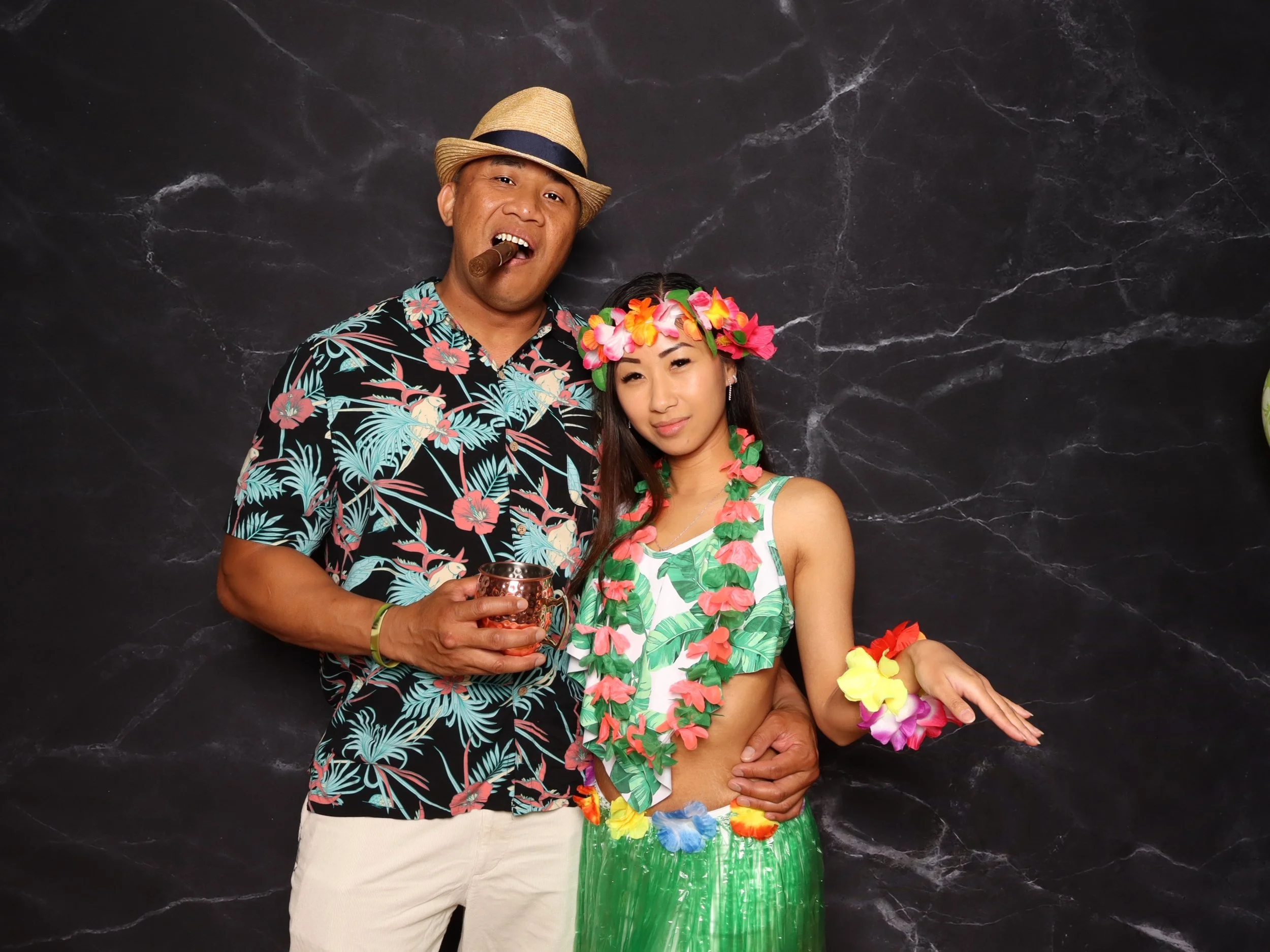 A man and a woman dressed in tropical themed outfits, with the man wearing a hawaiian shirt and the woman wearing a hula skirt and a flower lei, standing against a black marble wall background.