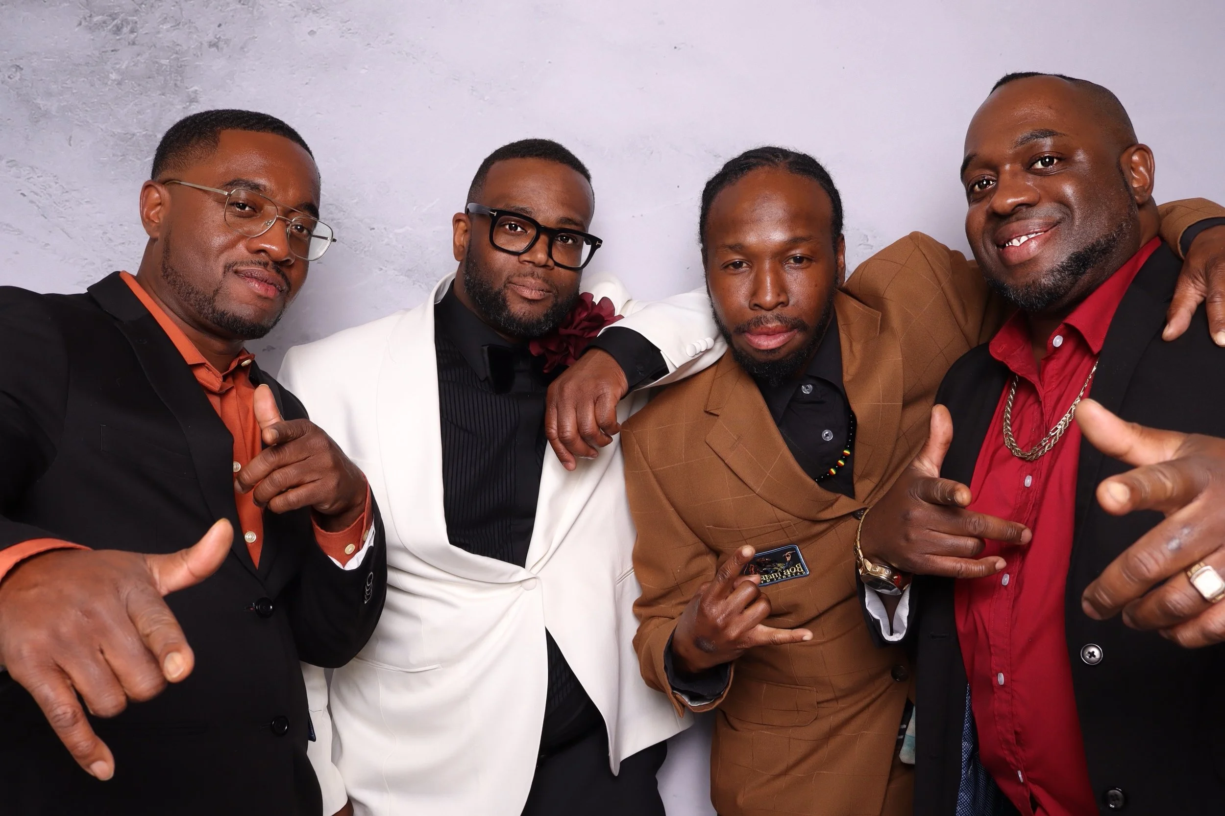 Four men dressed in formal and stylish attire posing together against a plain wall, pointing and making gestures.