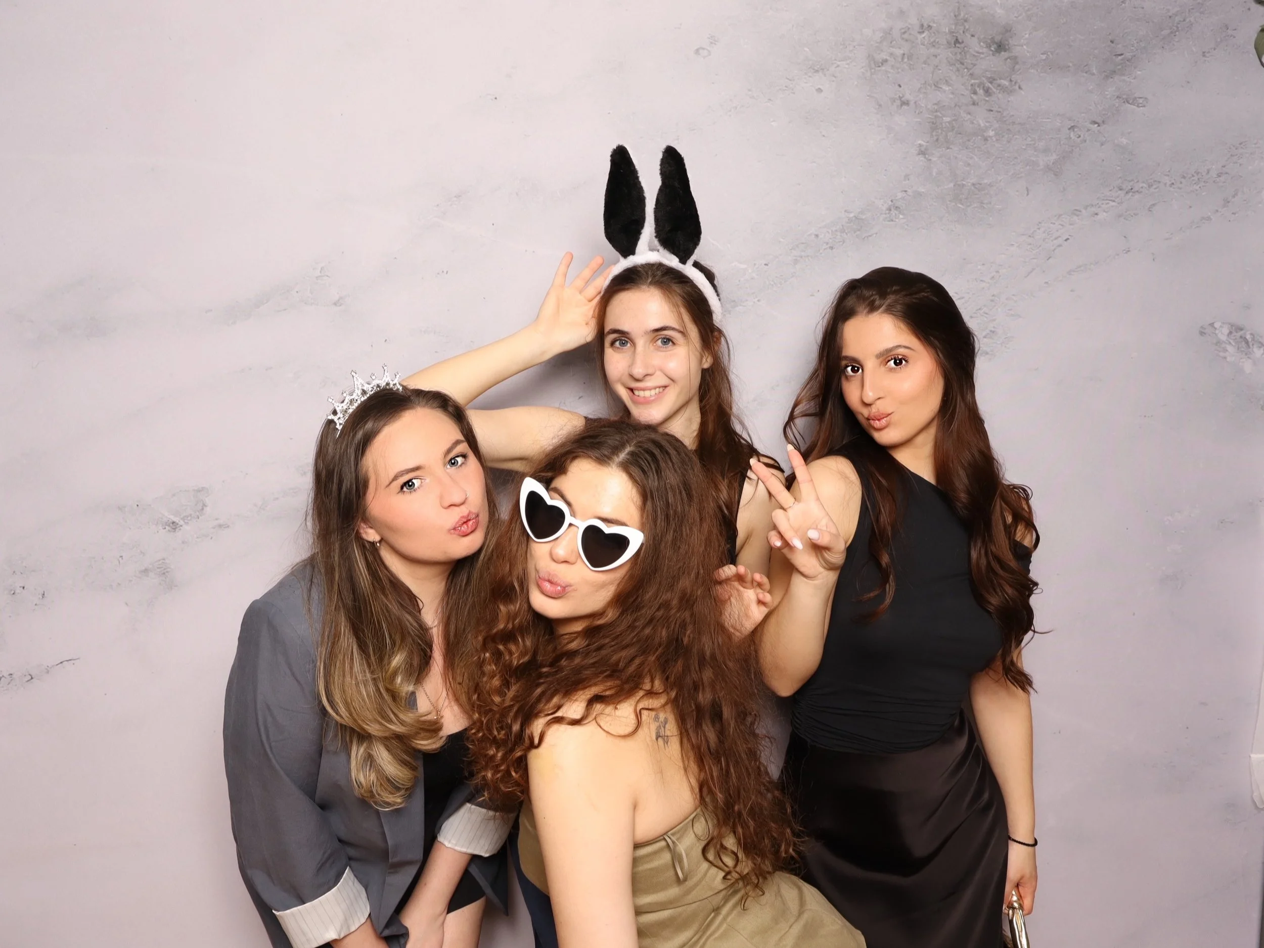Four young women posing with playful expressions and accessories, including a crown, sunglasses, bunny ears, and peace sign, against a plain background.
