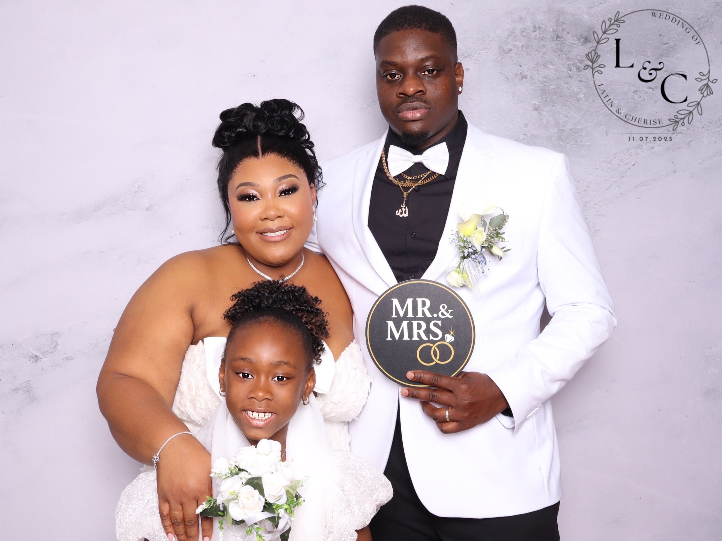 A family at a wedding, with a woman, a young girl, and a man in formal attire. The woman and girl are dressed in white, and the man is wearing a white tuxedo with a black shirt and bow tie. The man holds a sign that says "MR. & MRS." and has wedding 