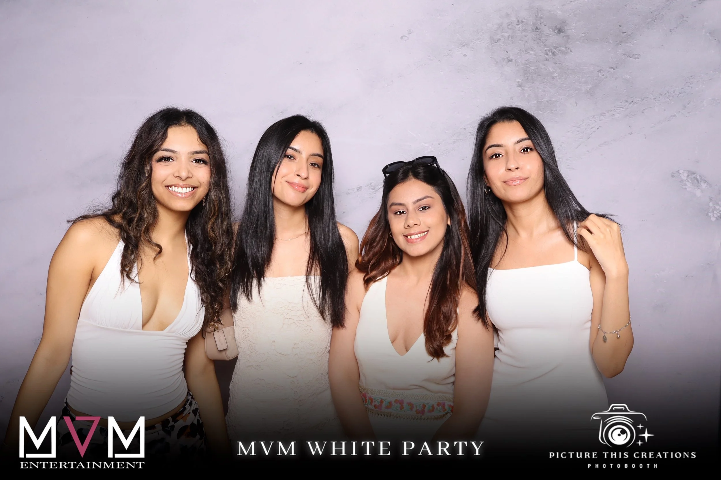 Four women in white tops smiling and posing for a photo at a party, standing against a gray textured backdrop with branding and event info at the bottom.