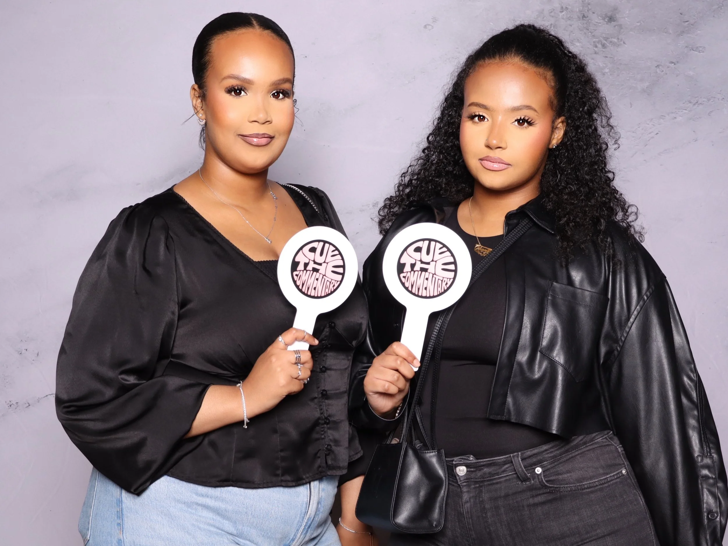 Two women standing side by side holding signs that read 'Cut The Comments', both wearing black outfits, one with a satin top and jeans, the other with a leather jacket and black pants, against a plain gray background.