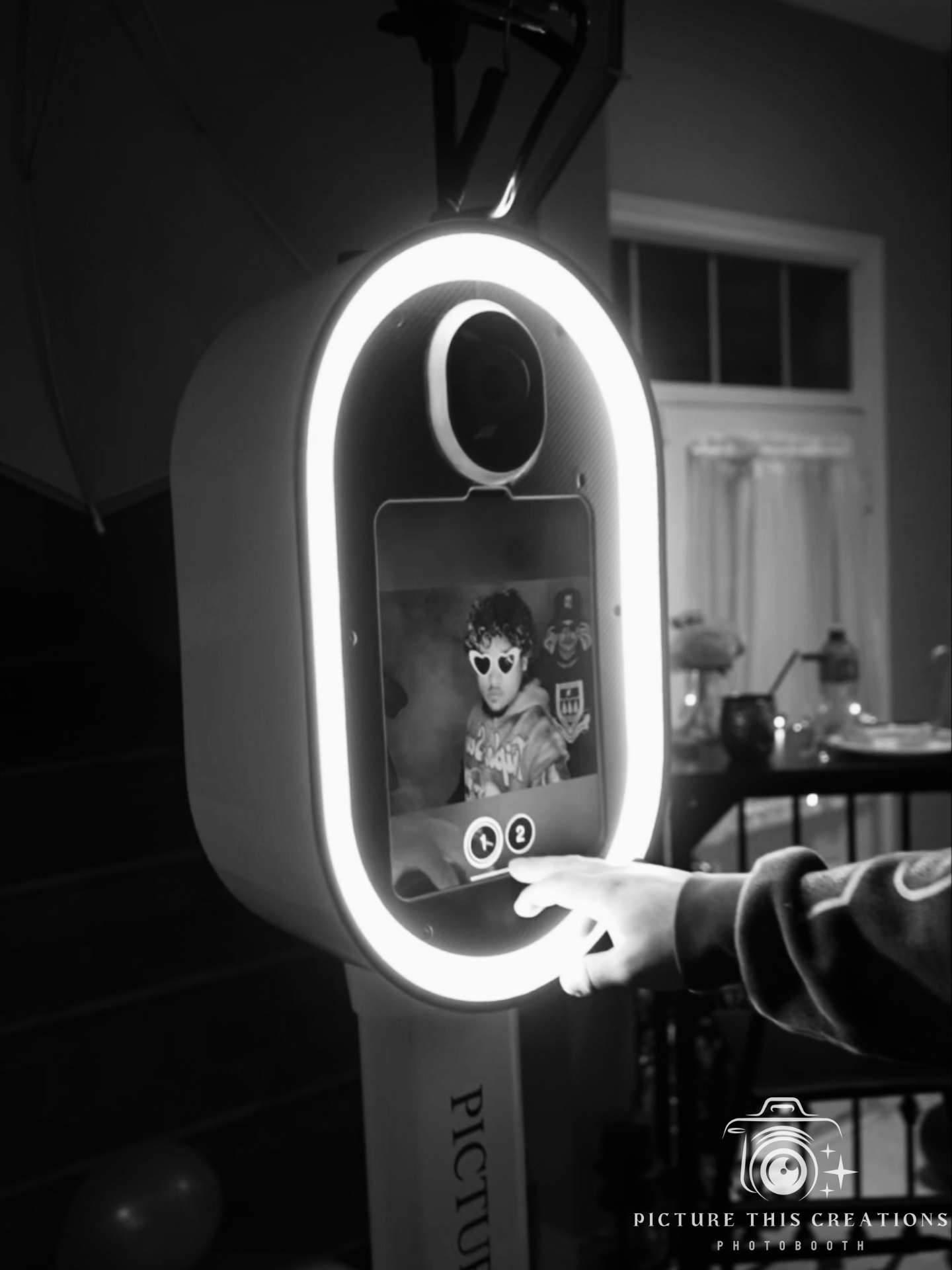 A person interacts with an illuminated photo booth, taking a picture of their reflection on the touchscreen display. The photo on the screen shows the person's face with heart-shaped glasses edited onto it.