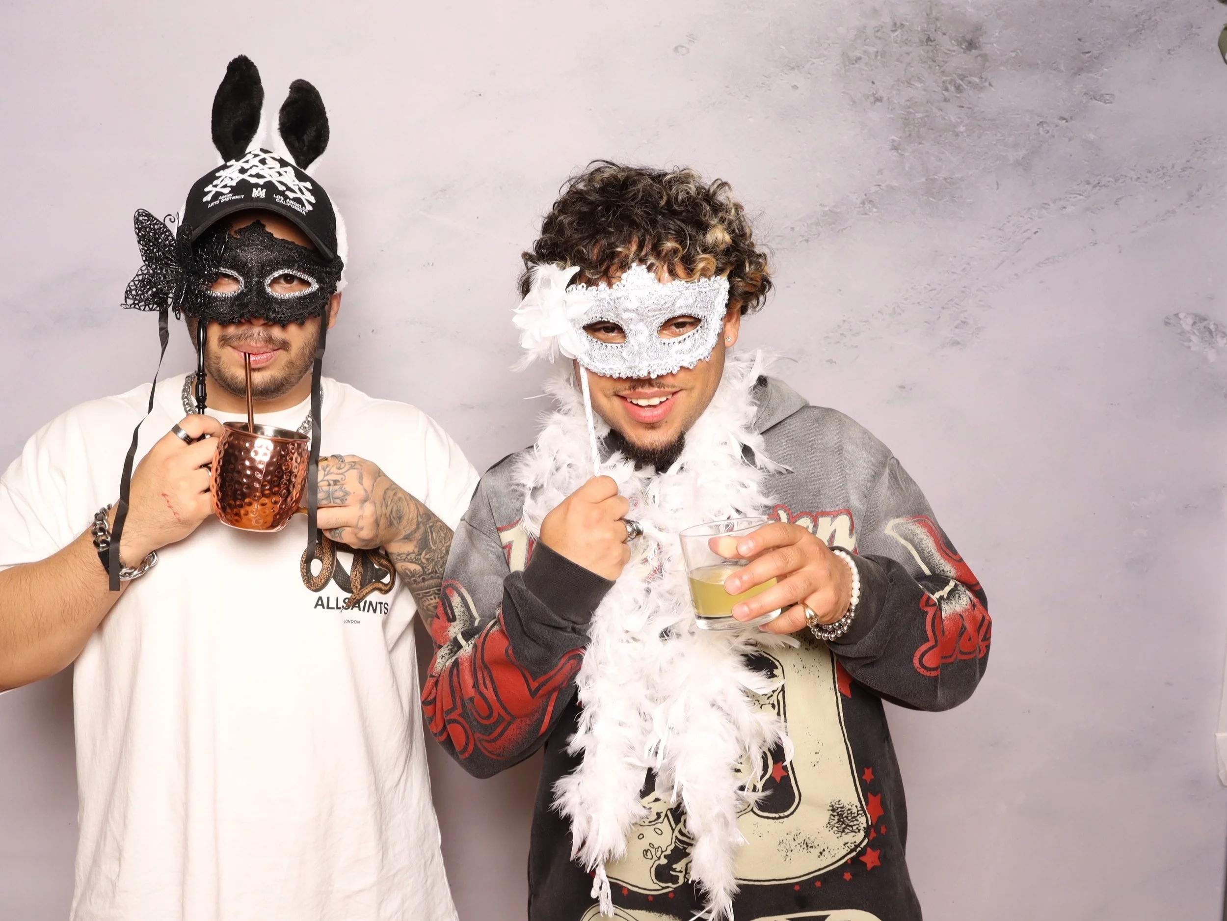 Two men wearing masks and accessories pose with drinks against a plain light gray background. The man on the left wears a black bunny mask with tall ears, a black cap, and a white t-shirt. The man on the right wears a white lace mask, a white feather