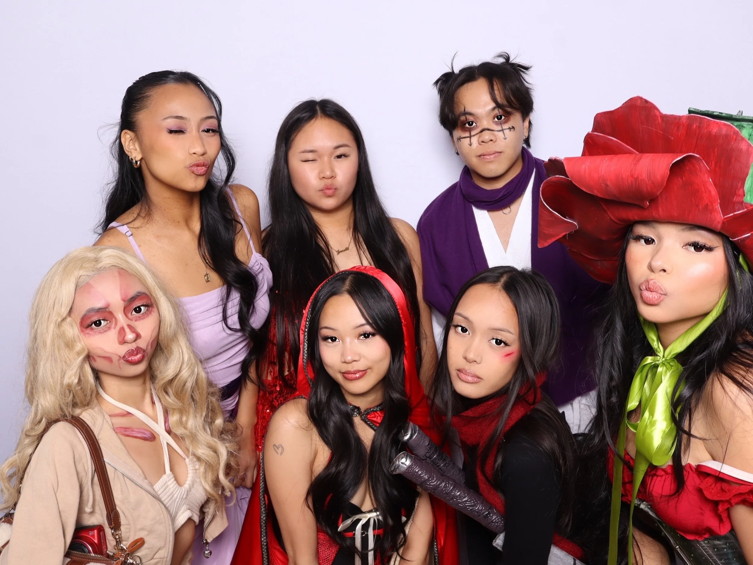 Group of people dressed in various colorful costumes, some with makeup and accessories, posing against a plain white background.