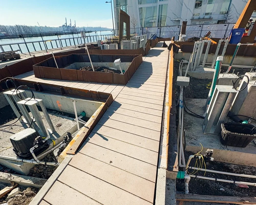 Construction site near waterfront with wood plank walkway, metal framework, and utility piping.