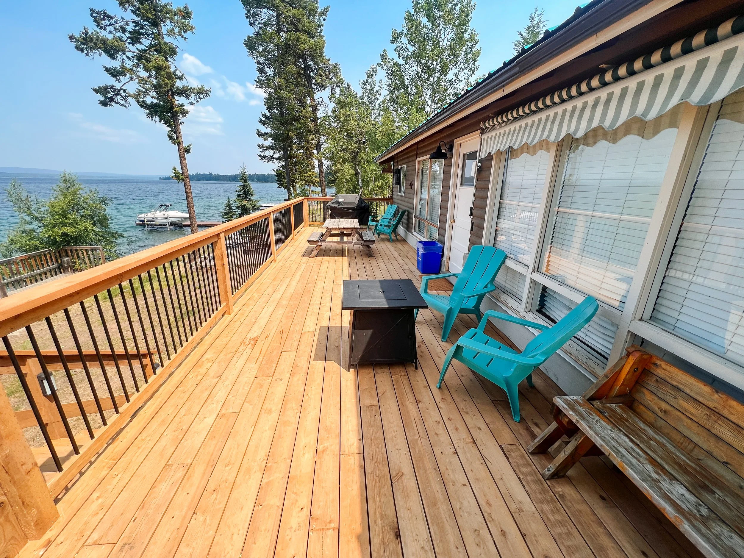 Wooden deck with turquoise chairs overlooking a lake, surrounded by trees. A boat is docked near the shoreline.