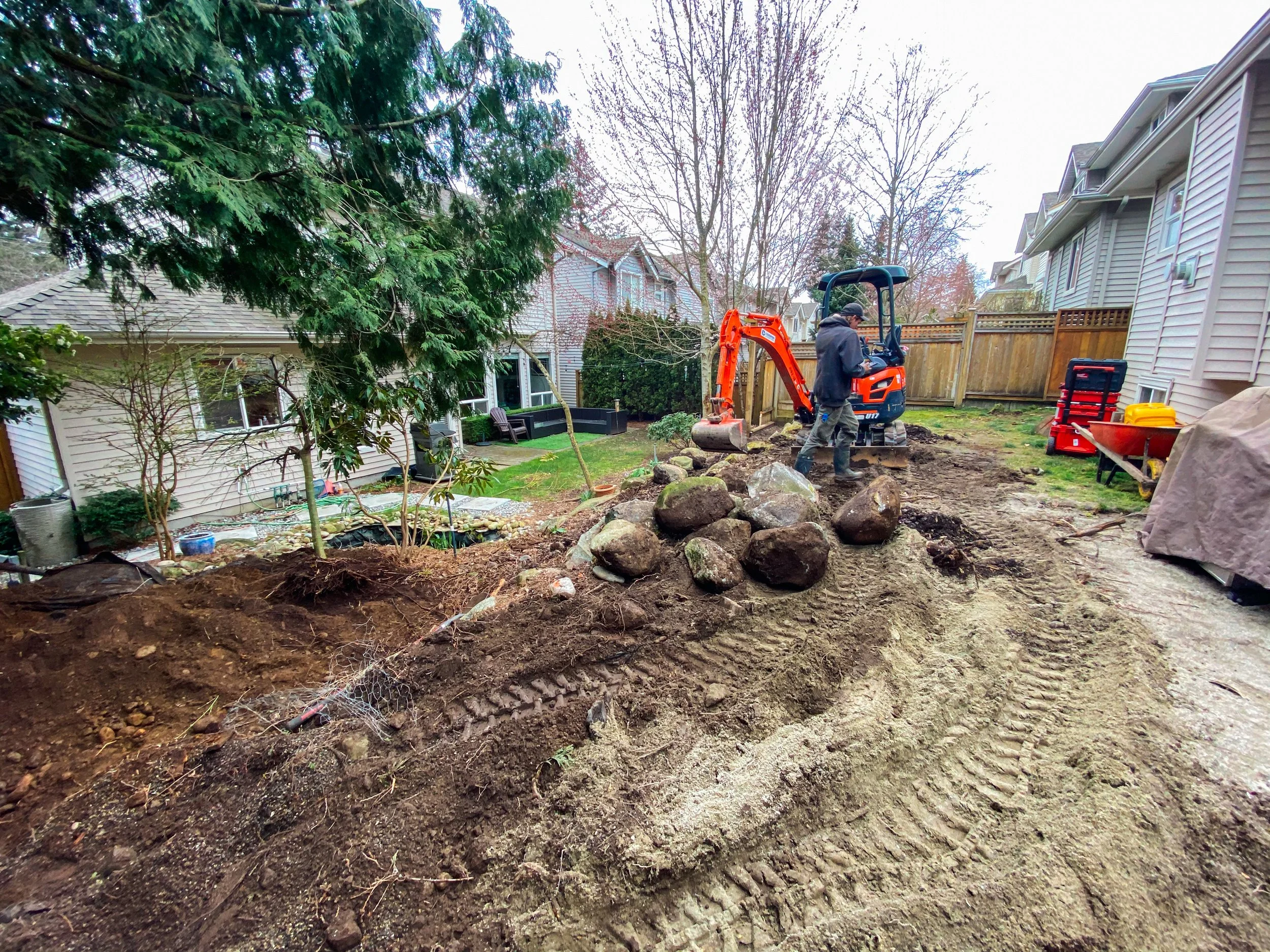 Person operating an orange excavator in a backyard, moving large rocks, with houses and trees in the background.