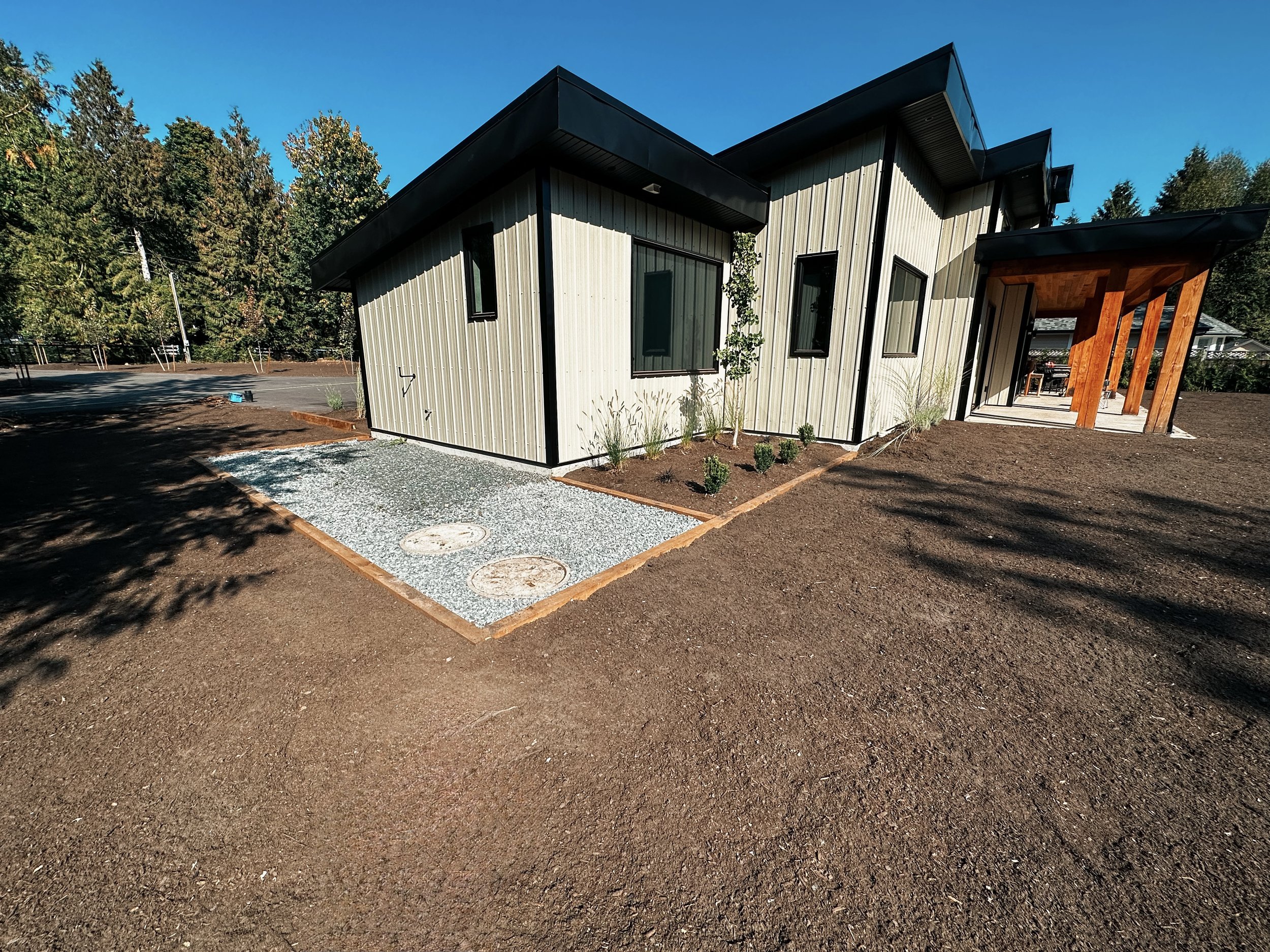 Modern house exterior with beige siding and black trim, surrounded by a freshly landscaped yard. A gravel area with circular covers is visible beside the house. Wooden architectural details are present, and the background features trees under a clear