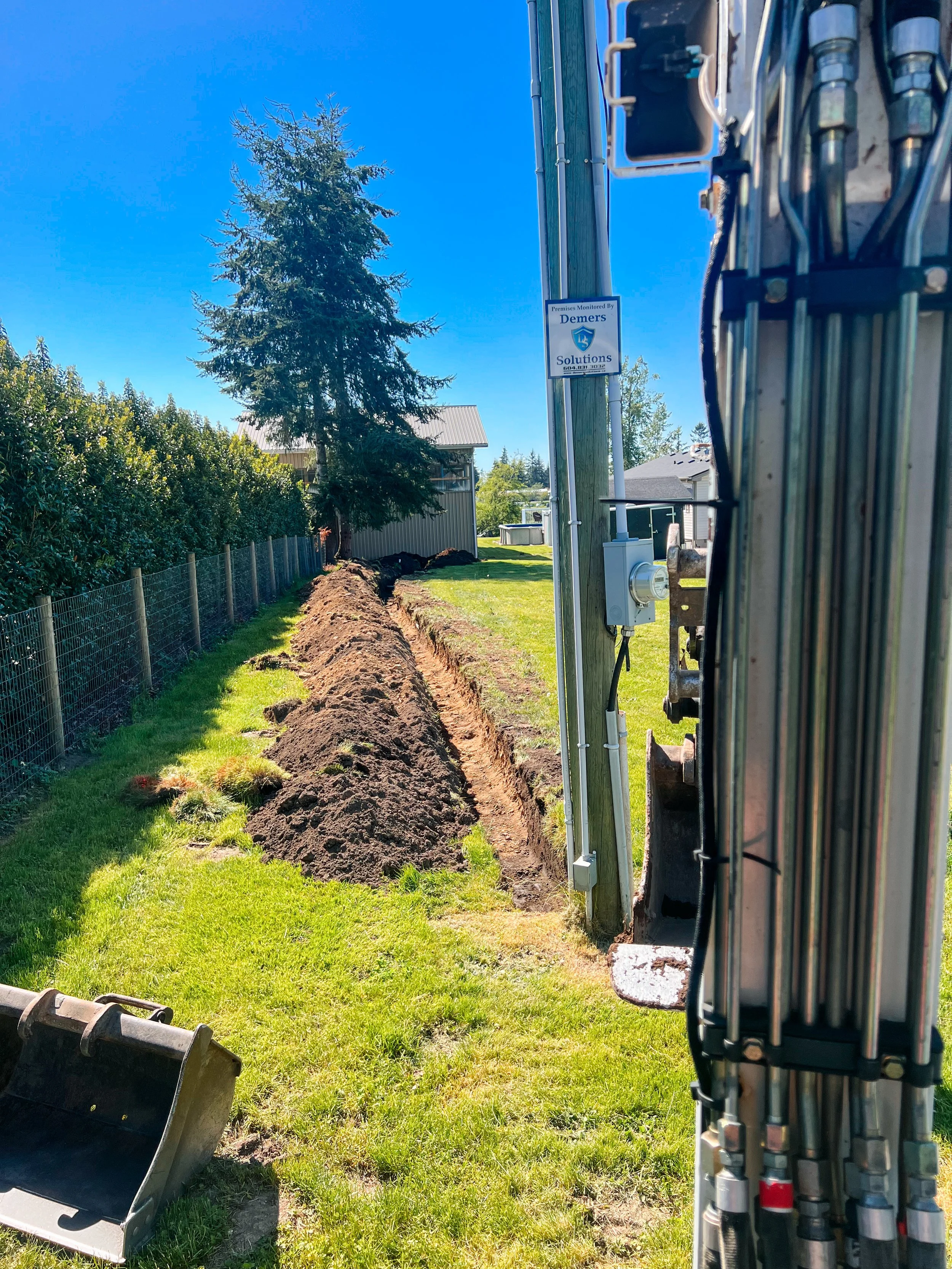 Excavation site with a long narrow trench, dirt piles, a tree, and construction equipment in a grassy area.