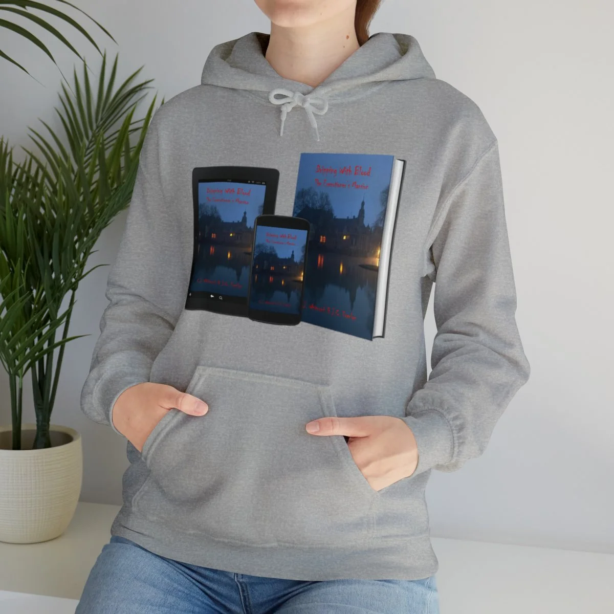 Person in a gray hoodie with a book cover on it, standing next to a potted plant against a plain wall.