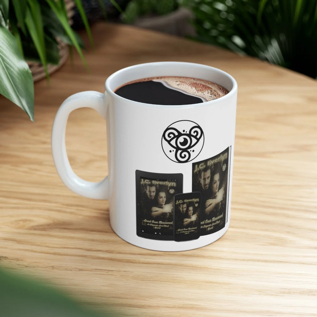 White coffee mug with a black tribal bear face design and images of a book cover, filled with black coffee, on a wooden table with green plants in the background.